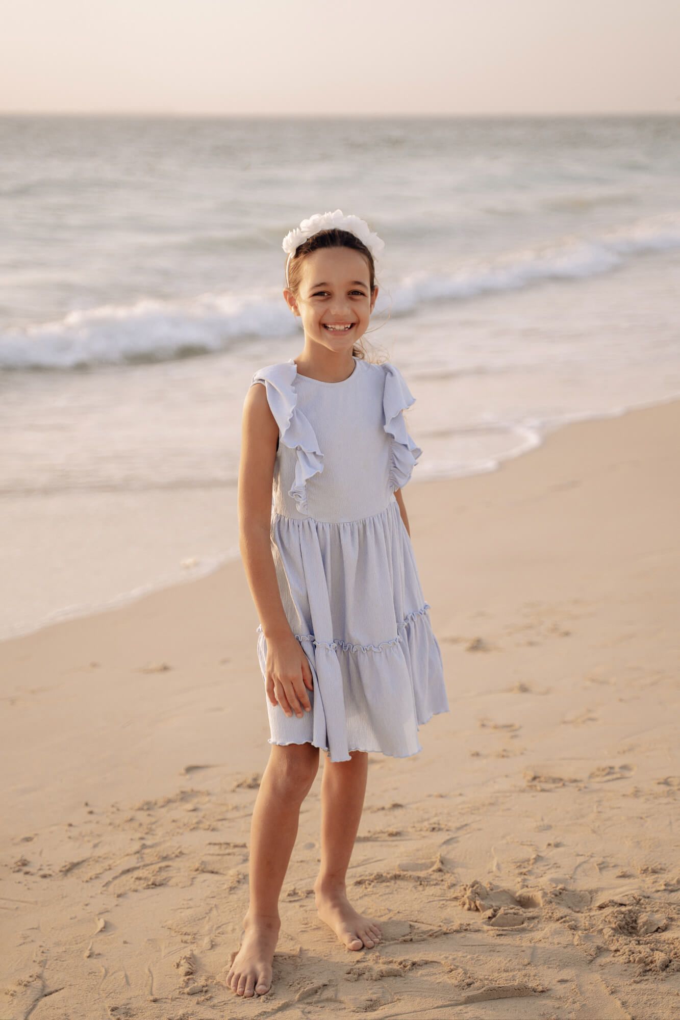 Child in a pale blue flutter sleeve dress runs and plays barefoot along the beach shoreline.