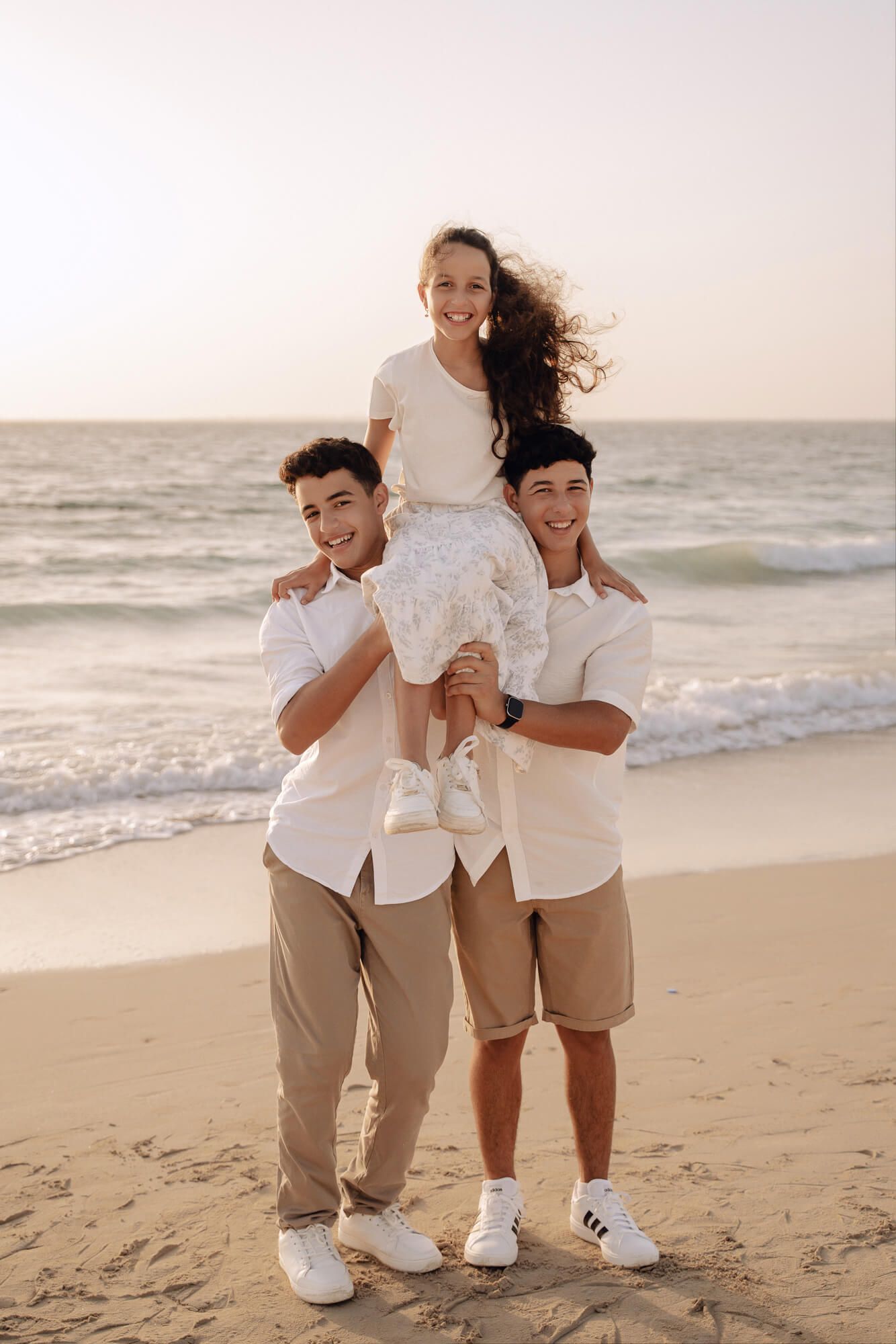 Three people in matching white shirts lift young girl on beach during golden hour.