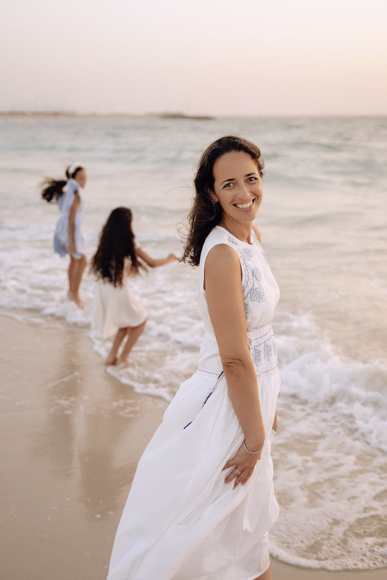 Woman in a flowing white dress dancing joyfully along the beach at sunset.
