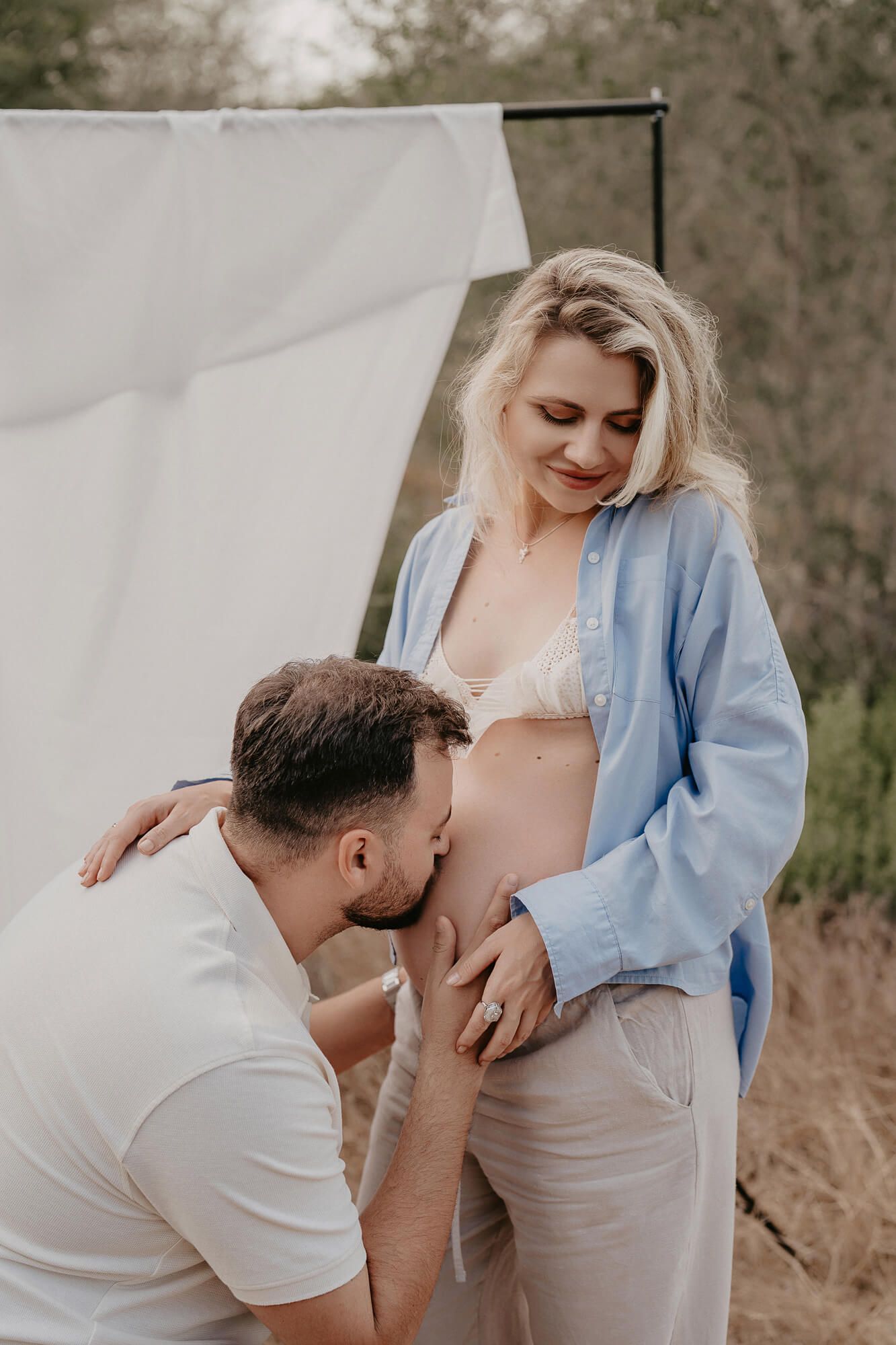 Intimate maternity photo showing couple in casual attire with light blue shirt and white clothing outdoors.