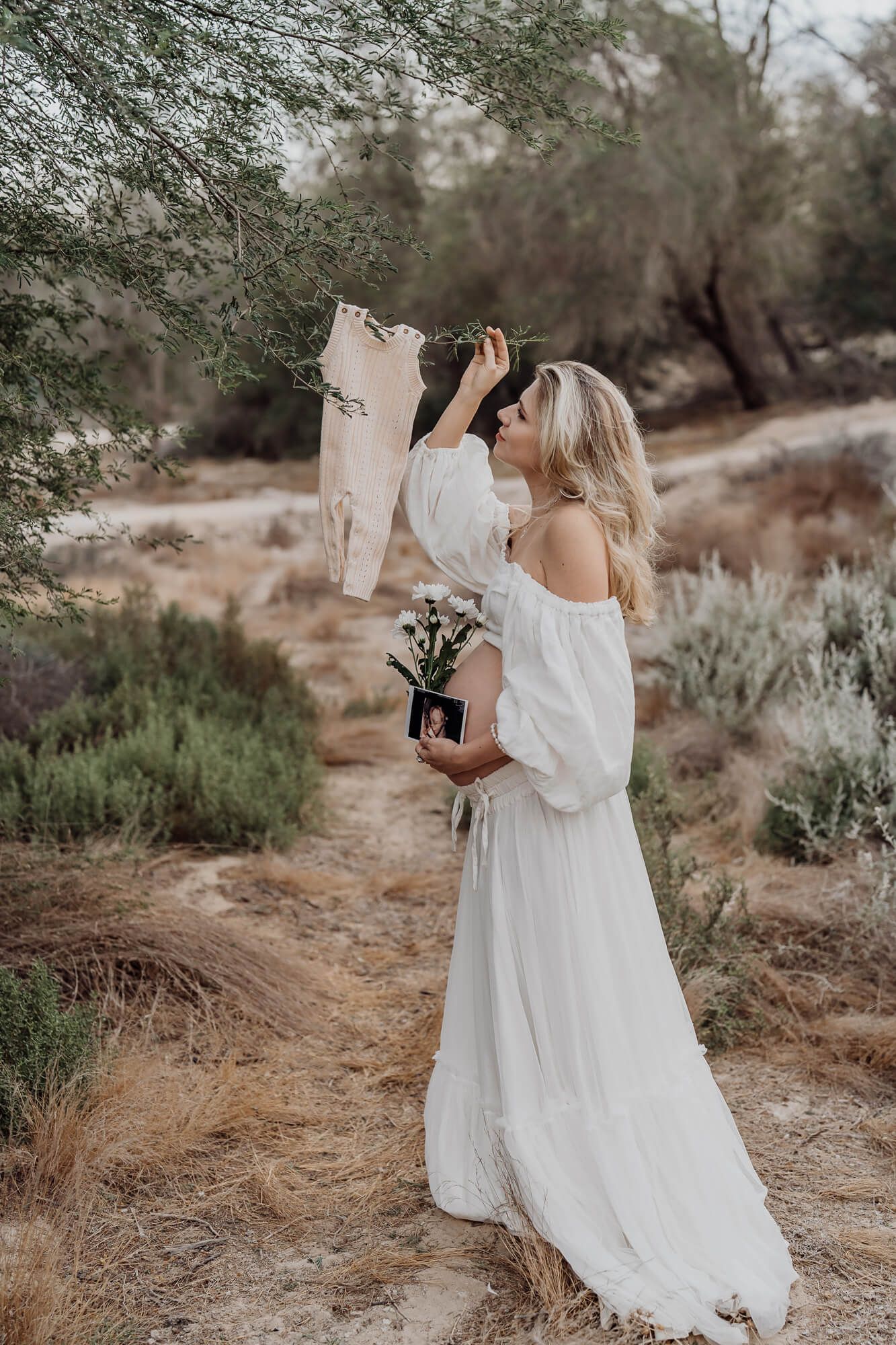 Woman in flowing white dress holding flowers and baby clothes in desert landscape.