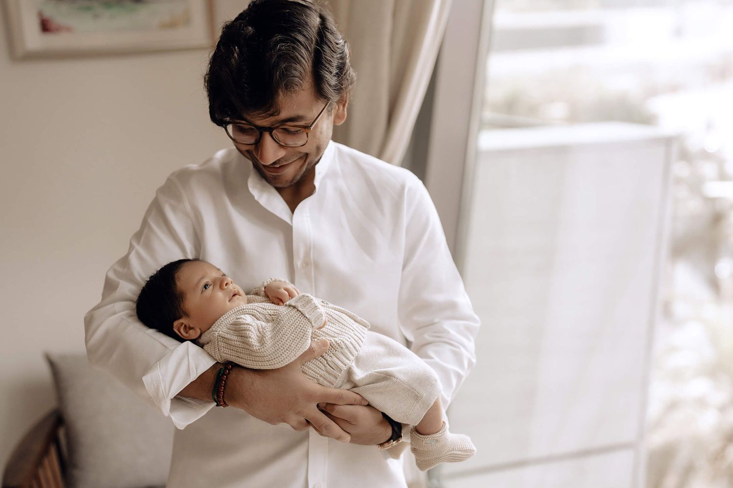 A person in a white shirt tenderly holds and gazes at a sleeping infant in a bright, sunlit room.