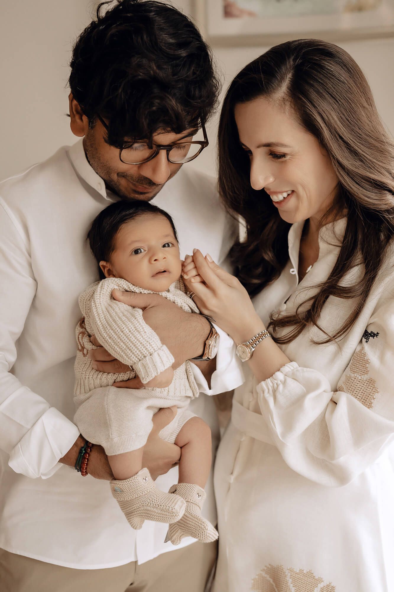 A tender family photo series in neutral tones shows parents sharing loving moments with their baby in matching white outfits.