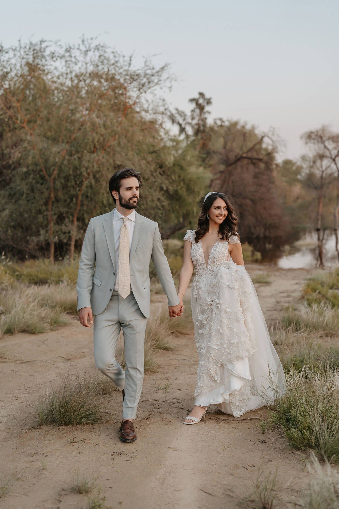 A couple in wedding attire walks together along a sandy beach path surrounded by autumn foliage and grasstops.