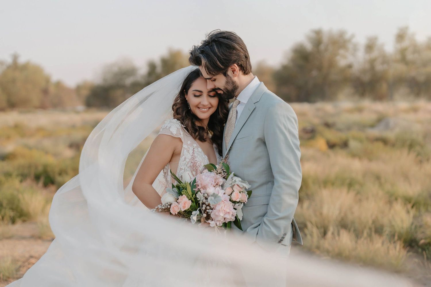 A couple takes romantic wedding photos in a desert setting with golden light and pink floral bouquet.