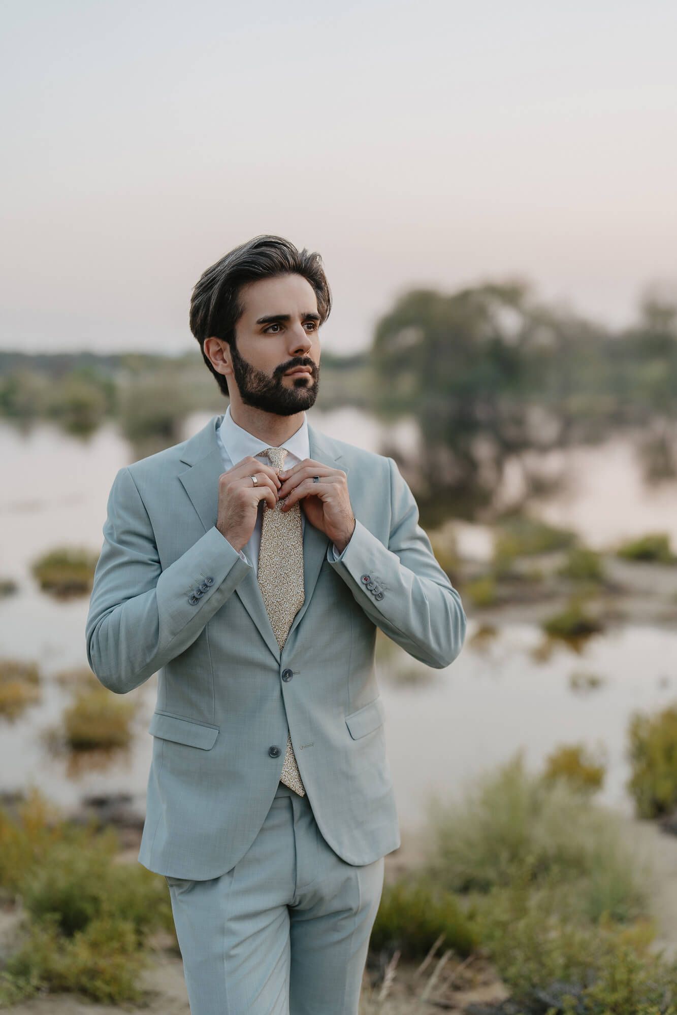 Well-dressed man in light blue suit adjusts his tie while standing in a serene lakeside setting at dusk.