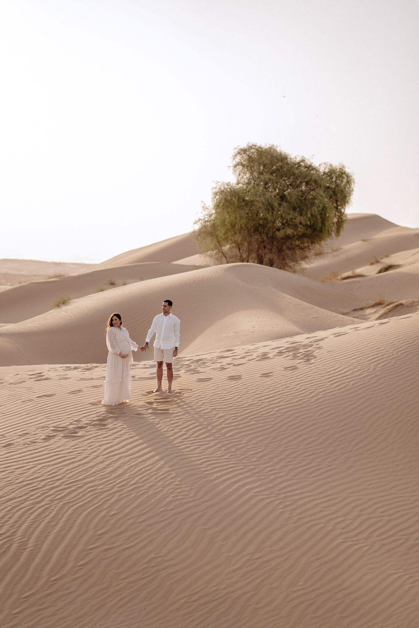 Two people in white attire stand together on golden sand dunes in a desert landscape with a lone tree in the background.