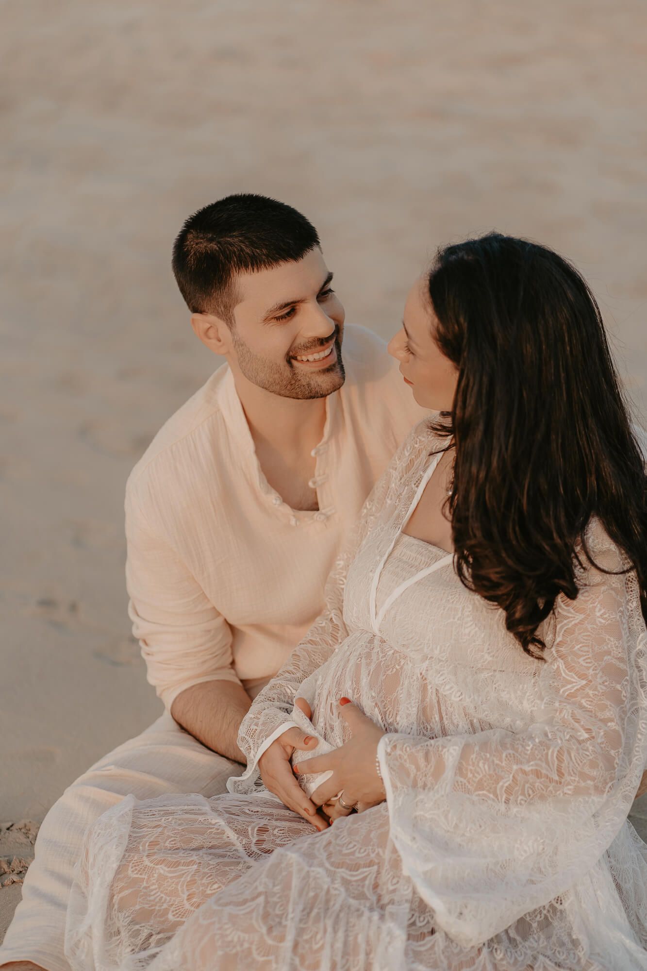 A romantic couple in light colored clothing shares an intimate candid moment together on sand.