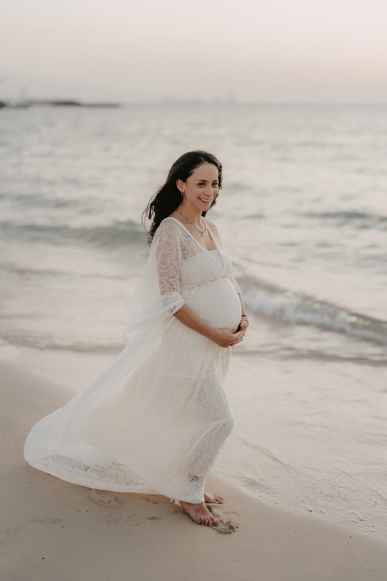 Expecting mother in flowing white lace dress walks barefoot along peaceful beach at sunset.
