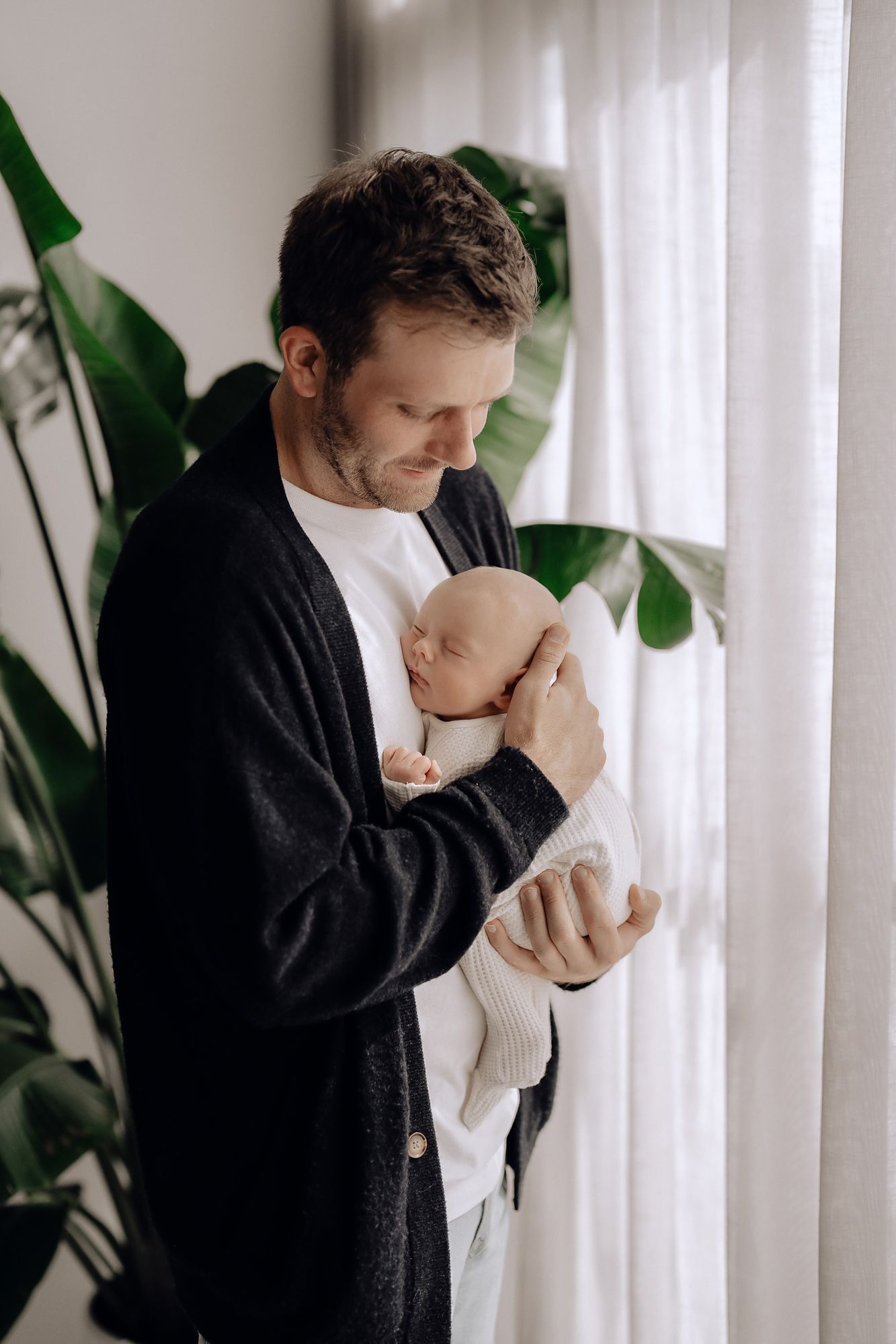 Tender moment captured as parent in black sweater embraces sleeping newborn baby near sheer white curtains and indoor plants.