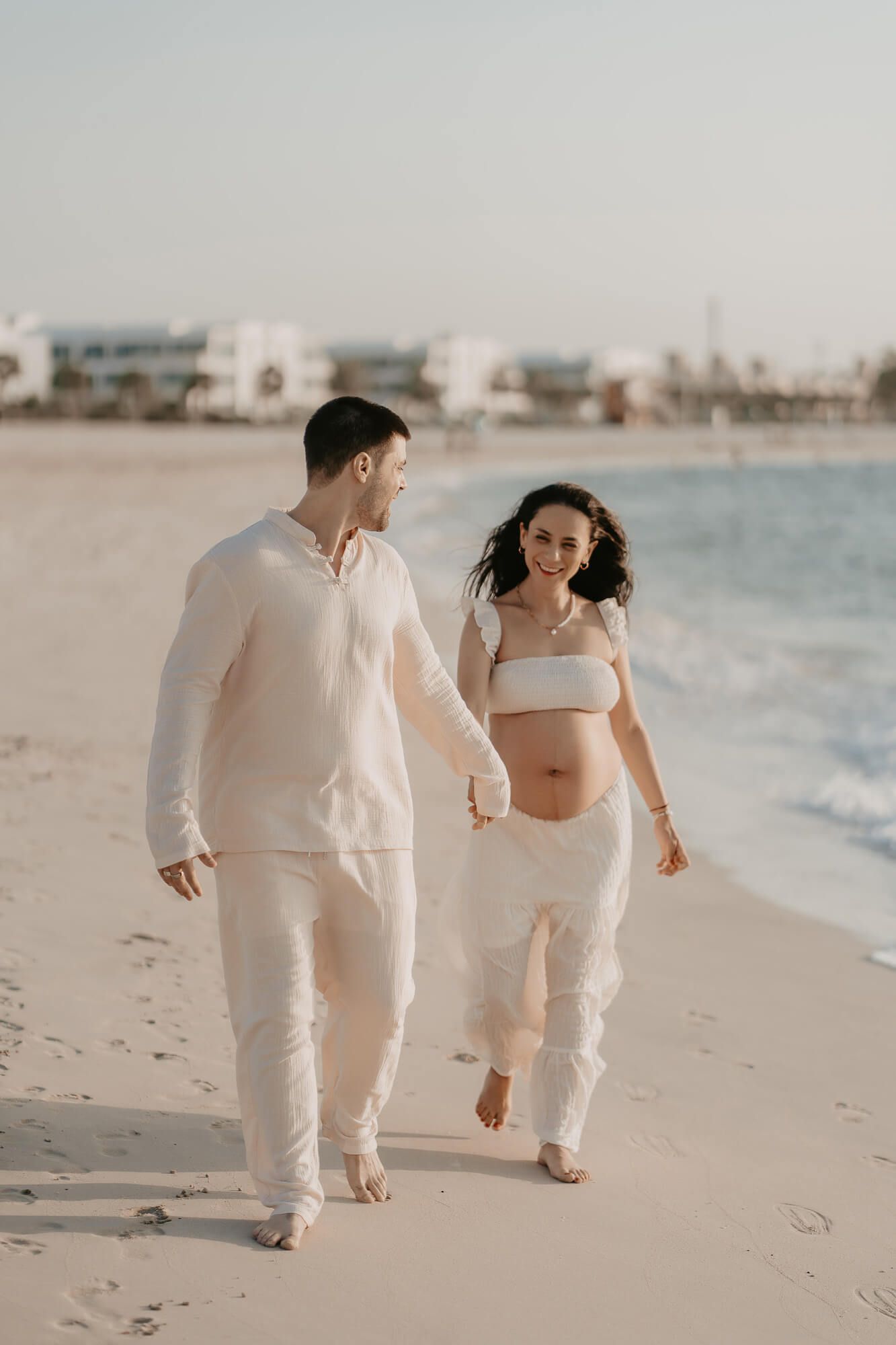 Couple in white outfits walking together on a sandy beach at sunset with ocean waves in the background.