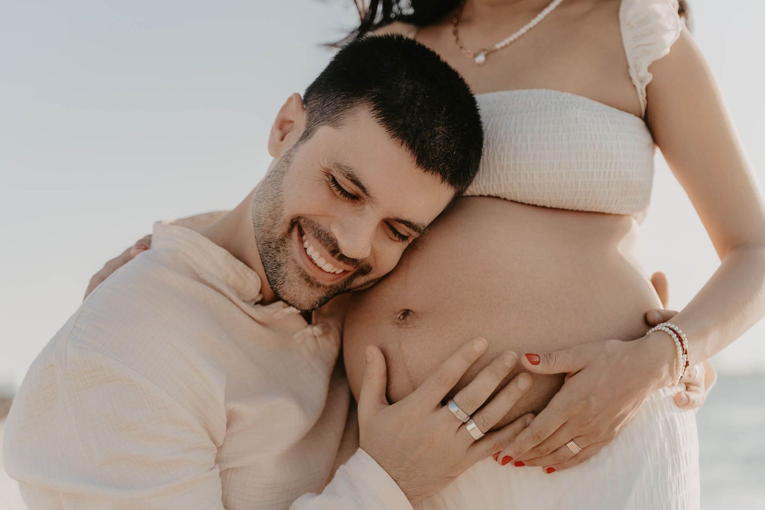 Intimate maternity photo showing loving embrace with pregnant belly in natural light against neutral background.