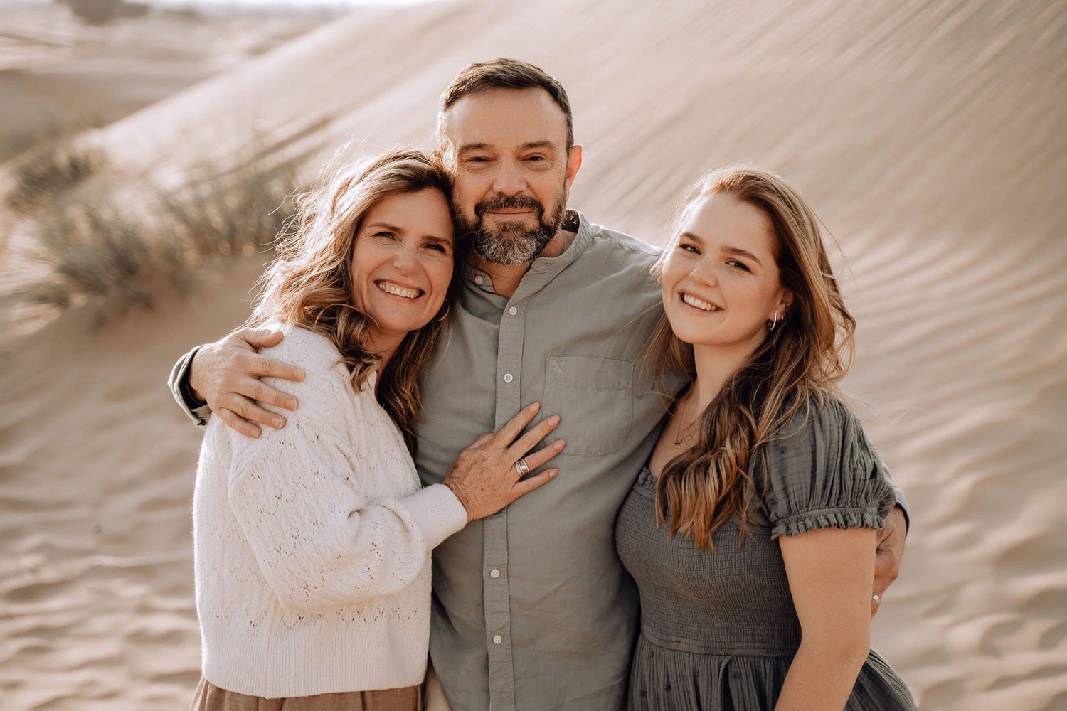 Three people smiling and embracing while posing for a portrait against sandy desert dunes.