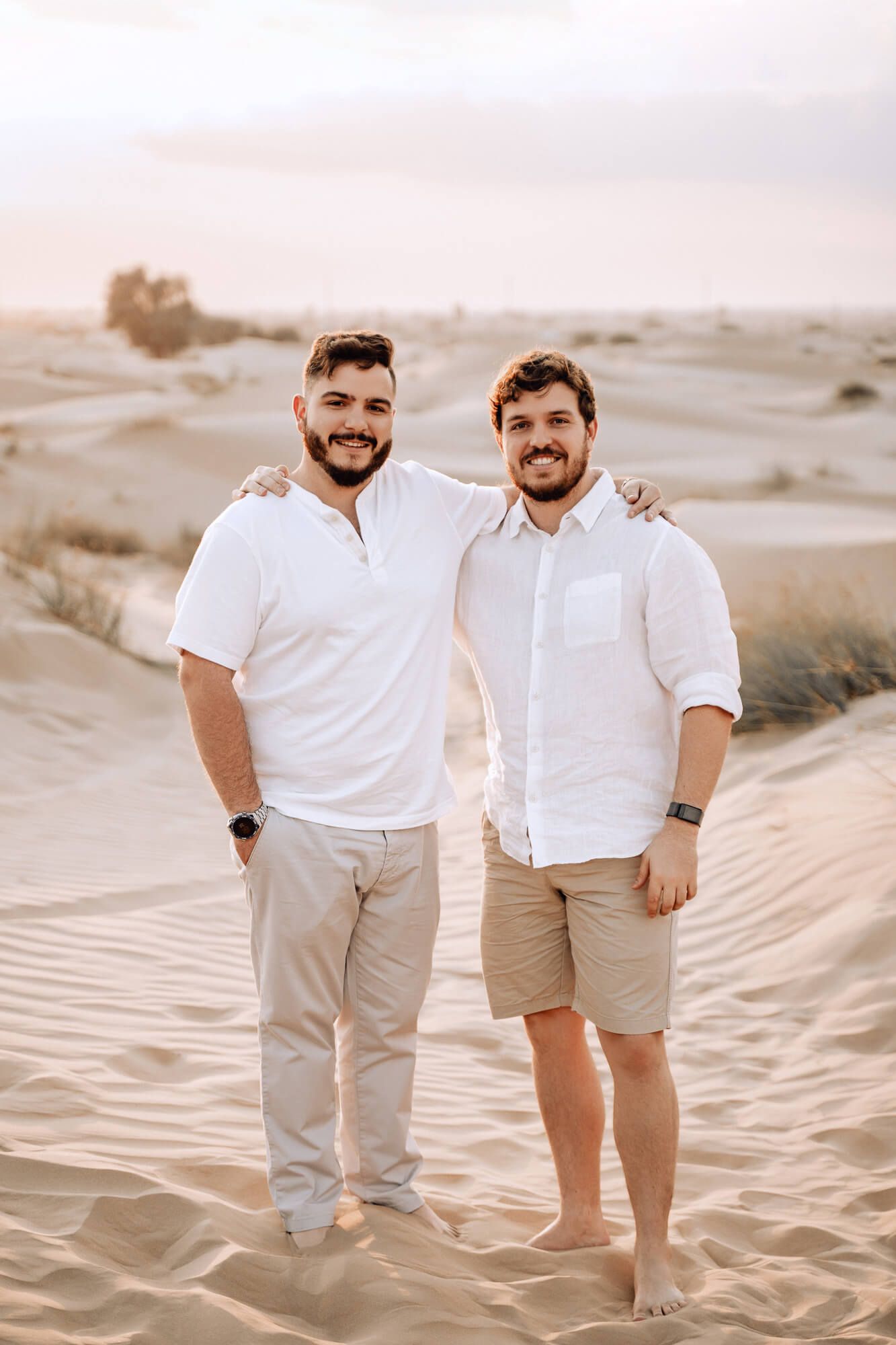 Two people wearing white shirts and khaki pants embrace while standing in sand dunes during golden hour.