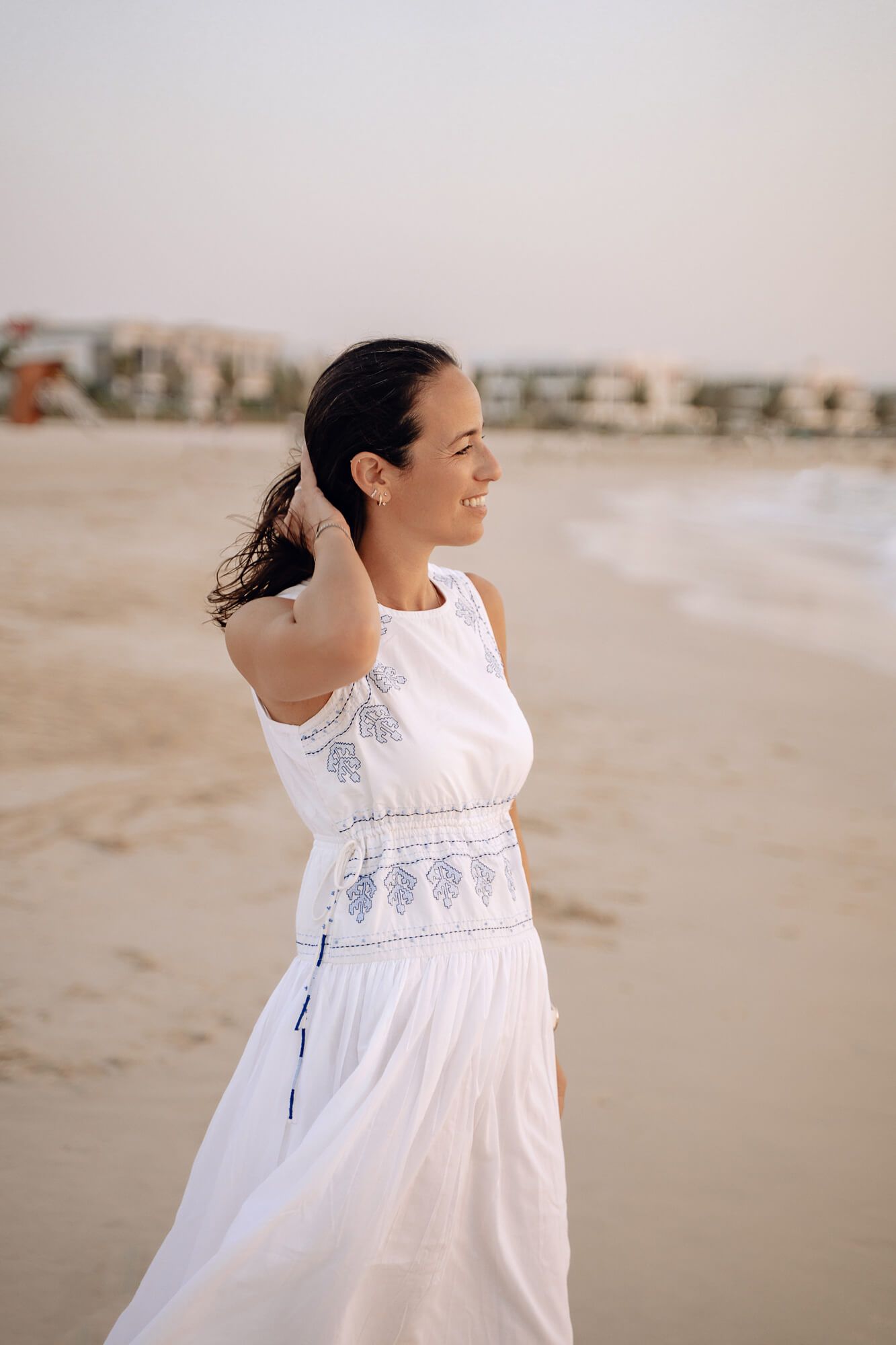 A white embroidered dress flows gracefully against the sandy beach backdrop.