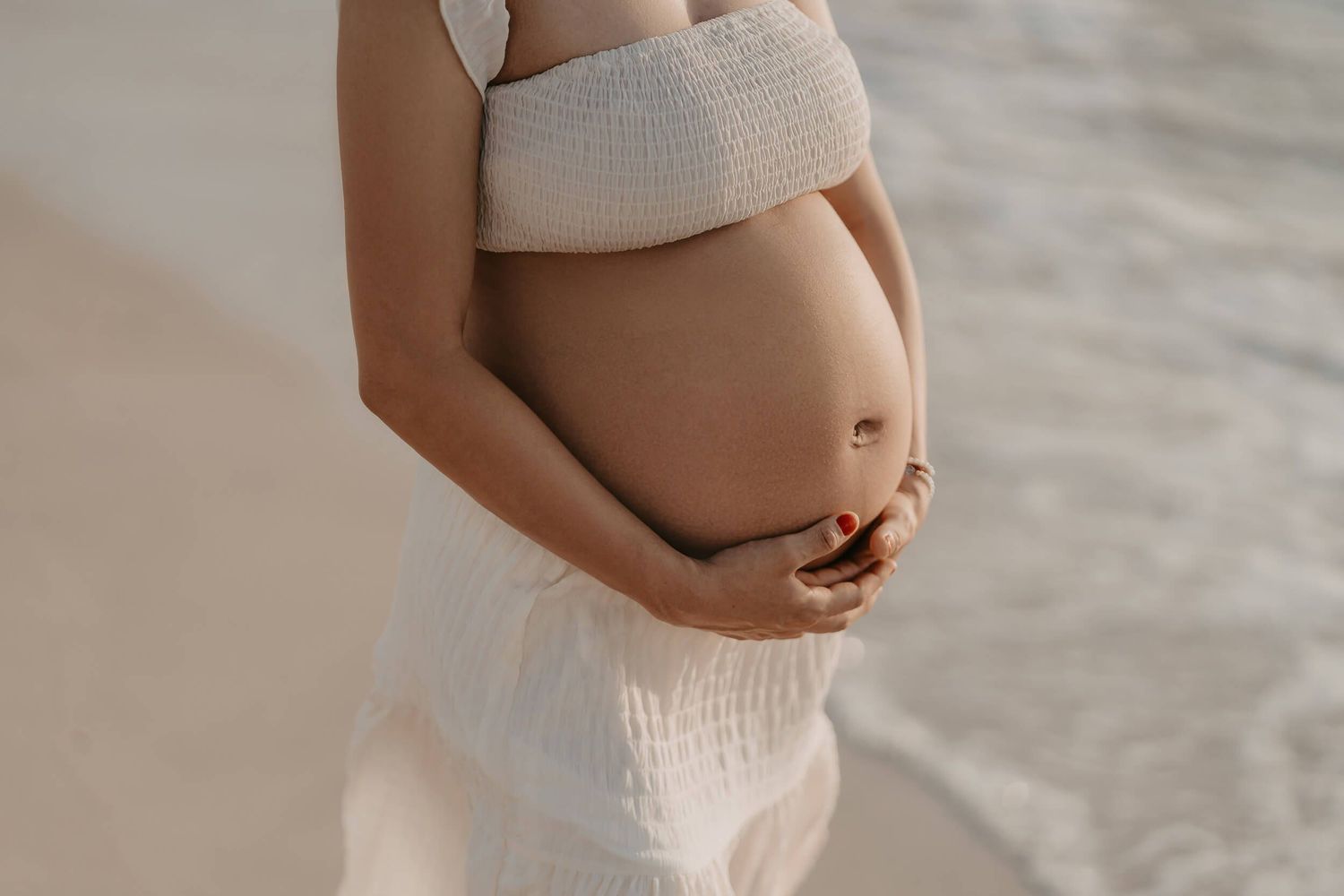 Close up of a pregnant belly in white fabric crop top and skirt against a neutral background.