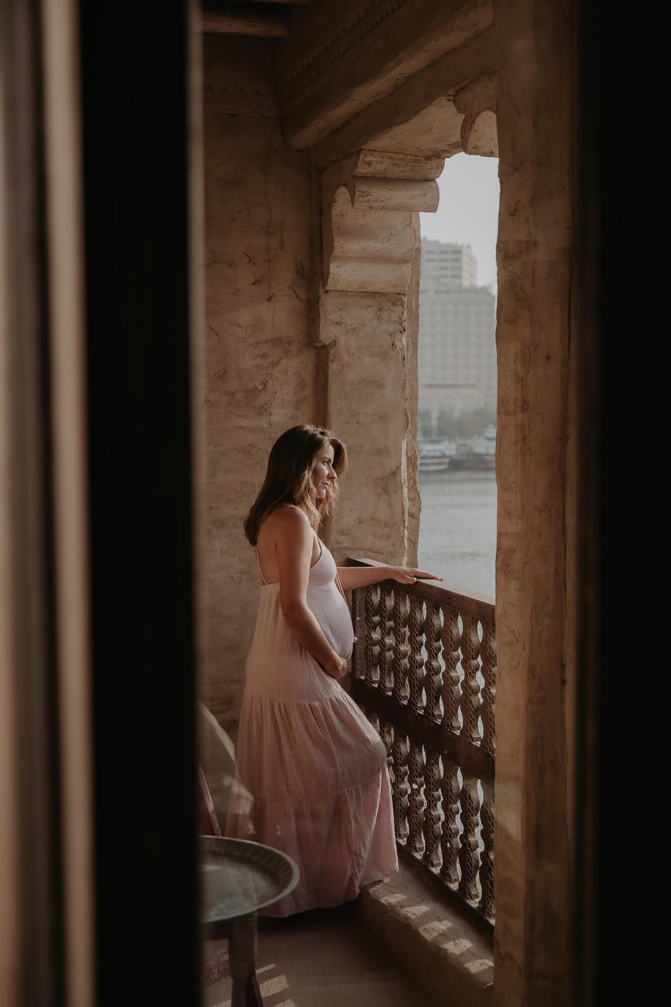 Woman in flowing pink dress stands on vintage stone balcony overlooking city waterfront in moody natural lighting.
