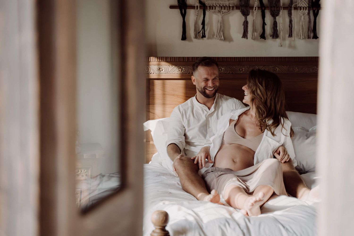 Family sits together on crisp white bedding in a bright, natural light-filled bedroom during intimate lifestyle photoshoot.