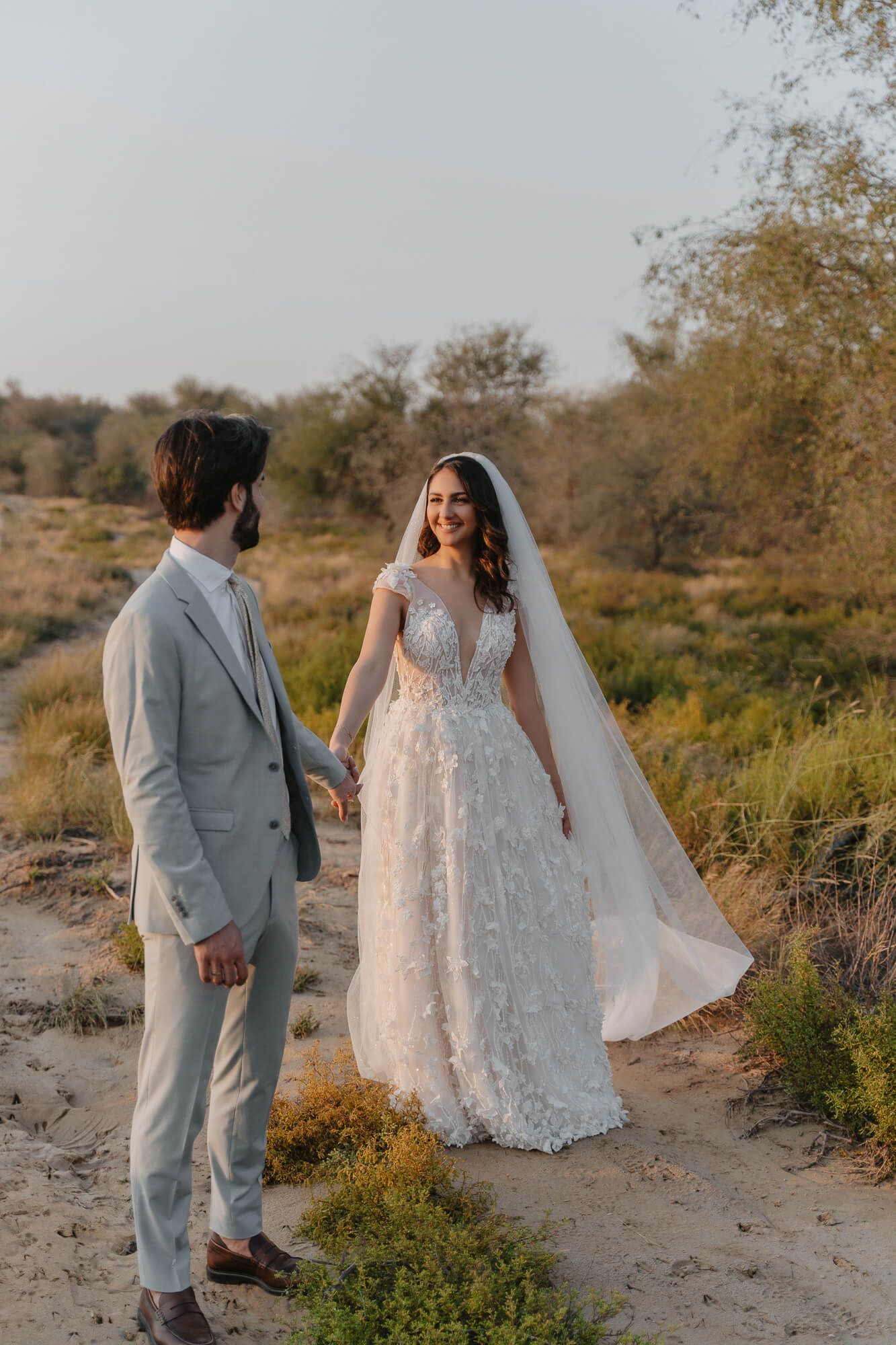 A couple in wedding attire pose together in a desert landscape during golden hour while wearing gray suit and lace gown.
