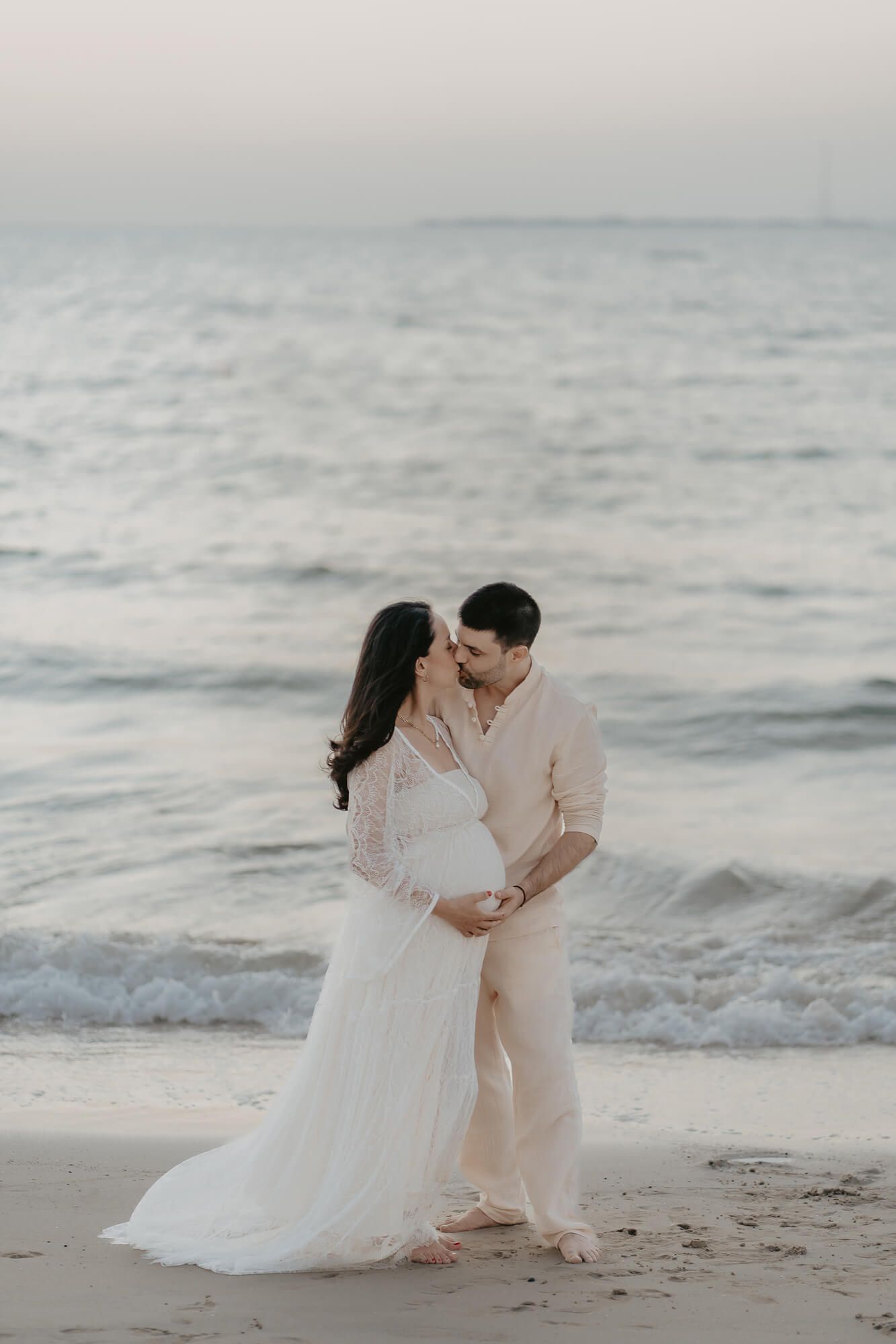 A romantic beach scene at sunset with a couple in light-colored attire embracing on the sandy shore.