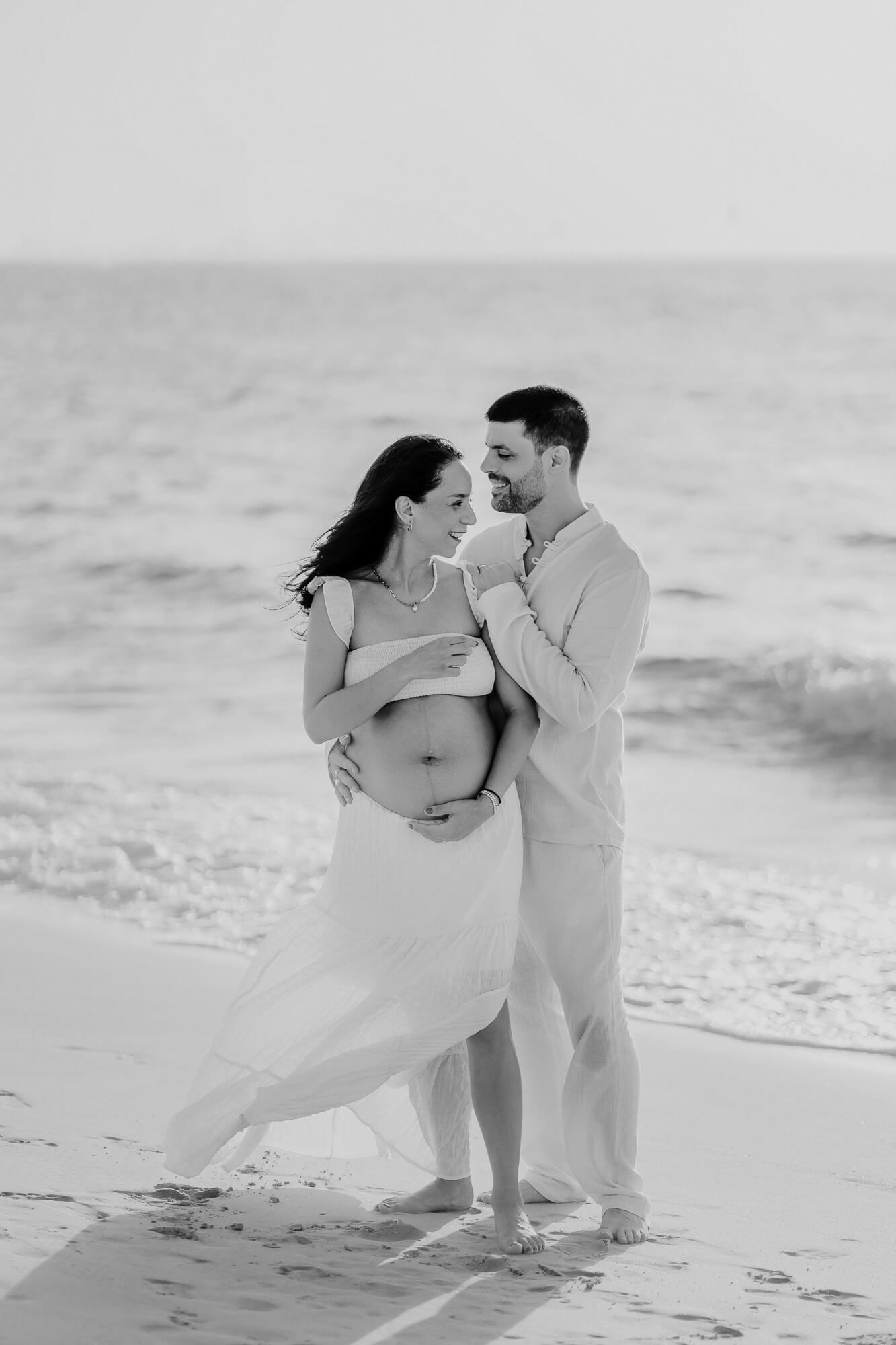 Silhouetted couple embraces on a sandy beach with ocean waves in the background, captured in black and white.