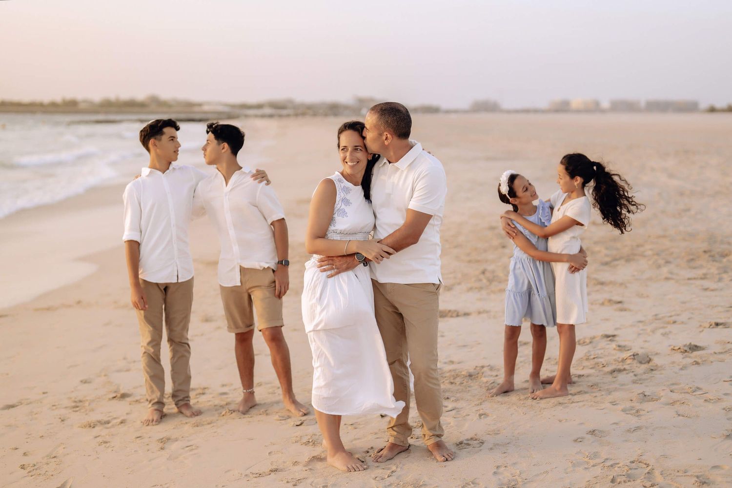 Family gathering at sunset on sandy beach wearing coordinated white and neutral outfits.