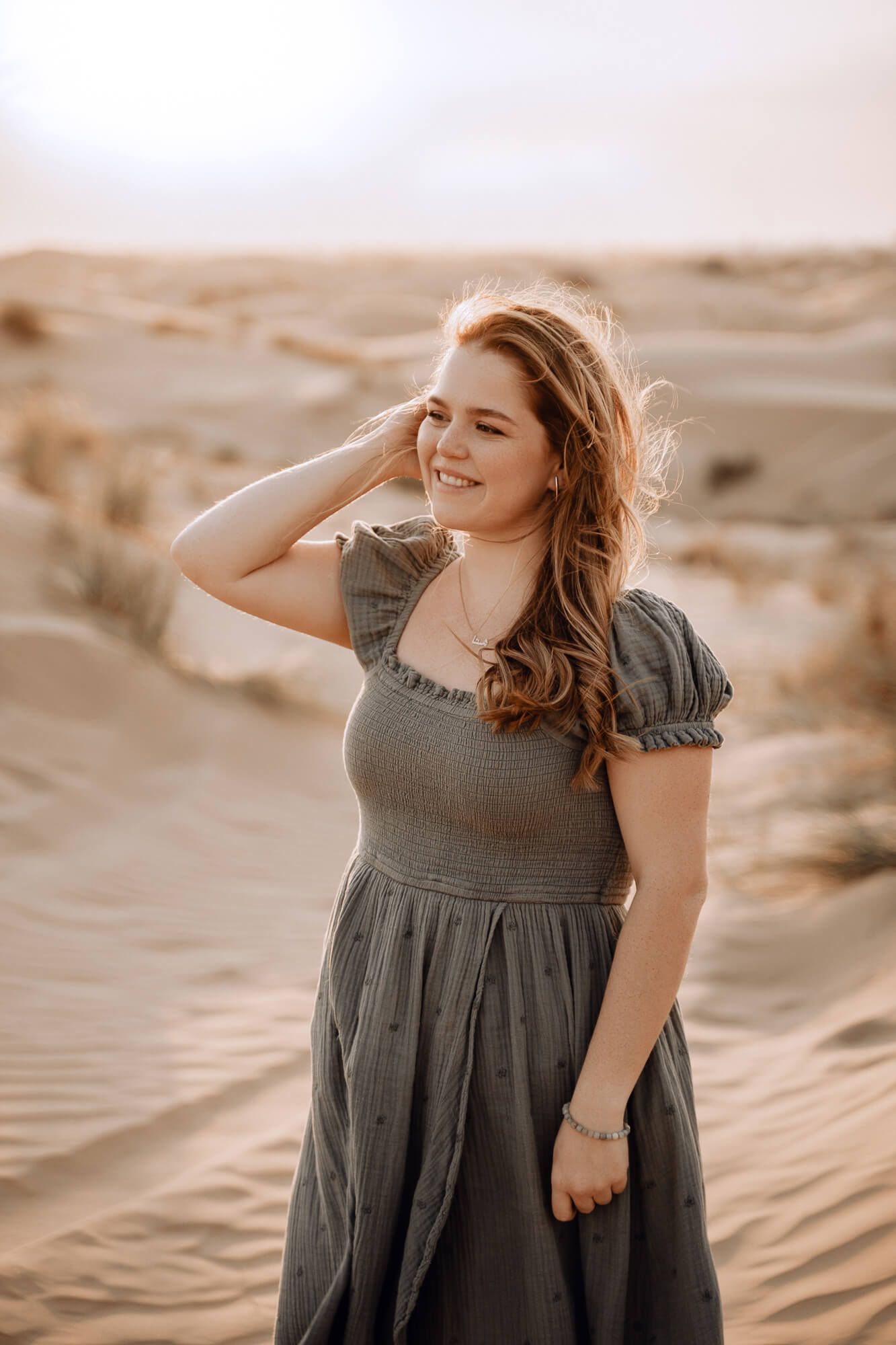 Woman in gray dress poses with windswept hair against desert landscape during golden hour.