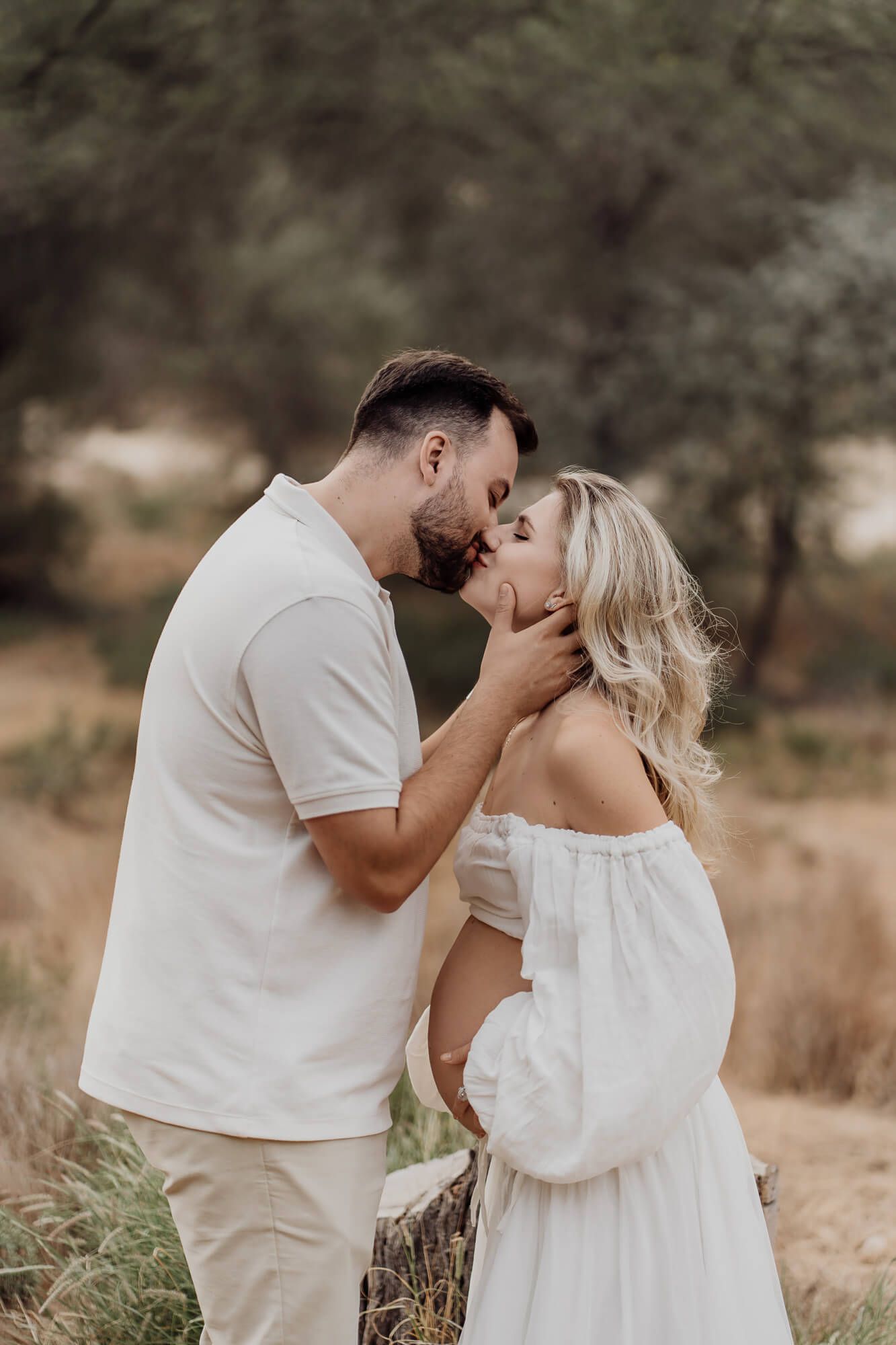 Romantic maternity photo of couple in white outfits sharing tender moment in nature.