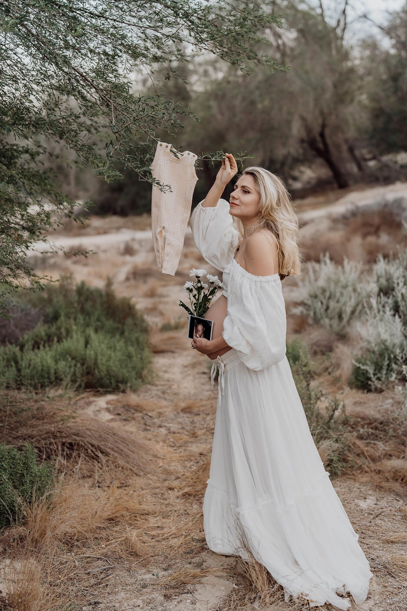 Woman in off-shoulder white dress holding infant clothes and flowers in desert setting.
