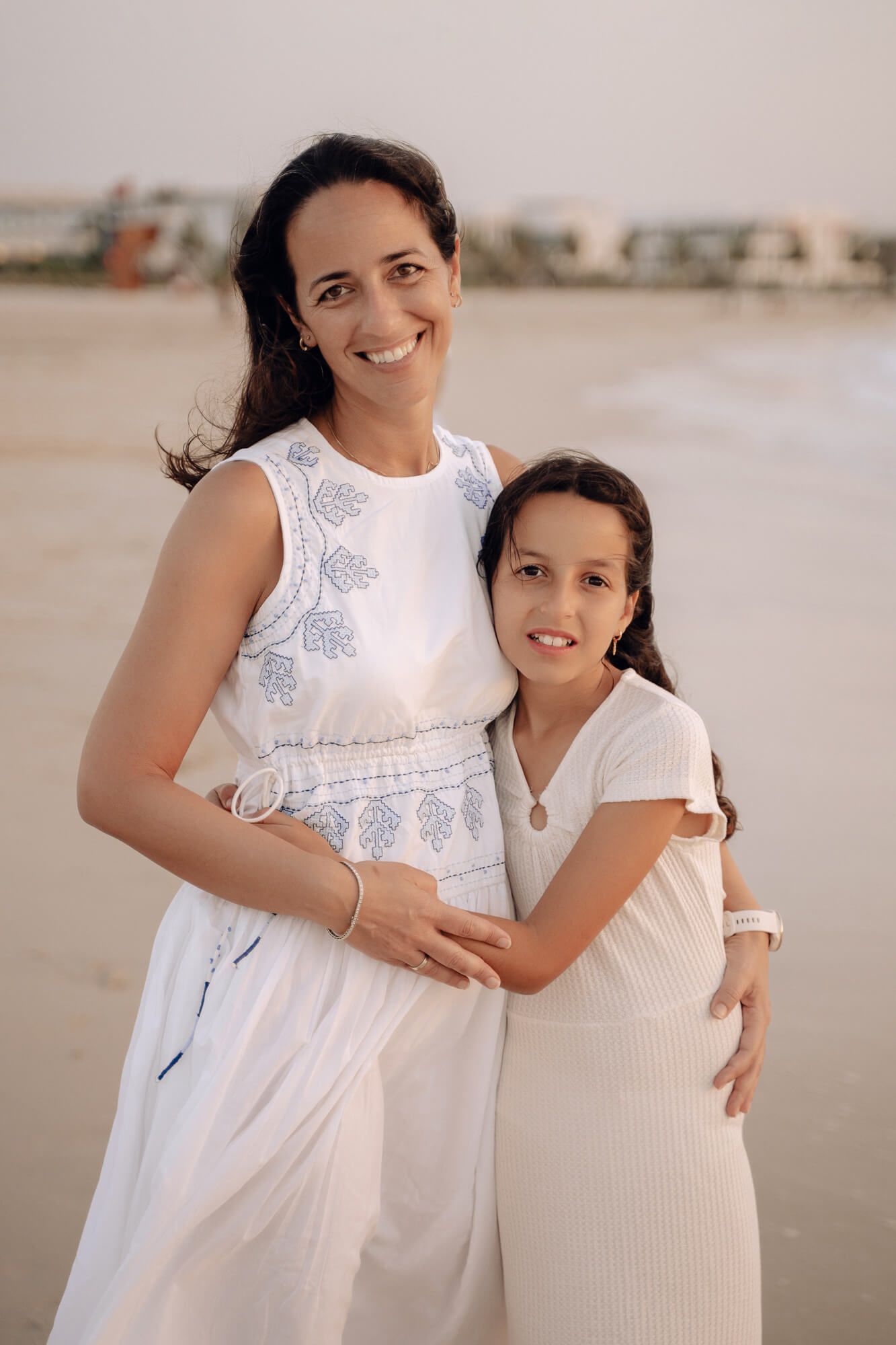 Two people in white outfits embrace warmly on a peaceful beach during golden hour.