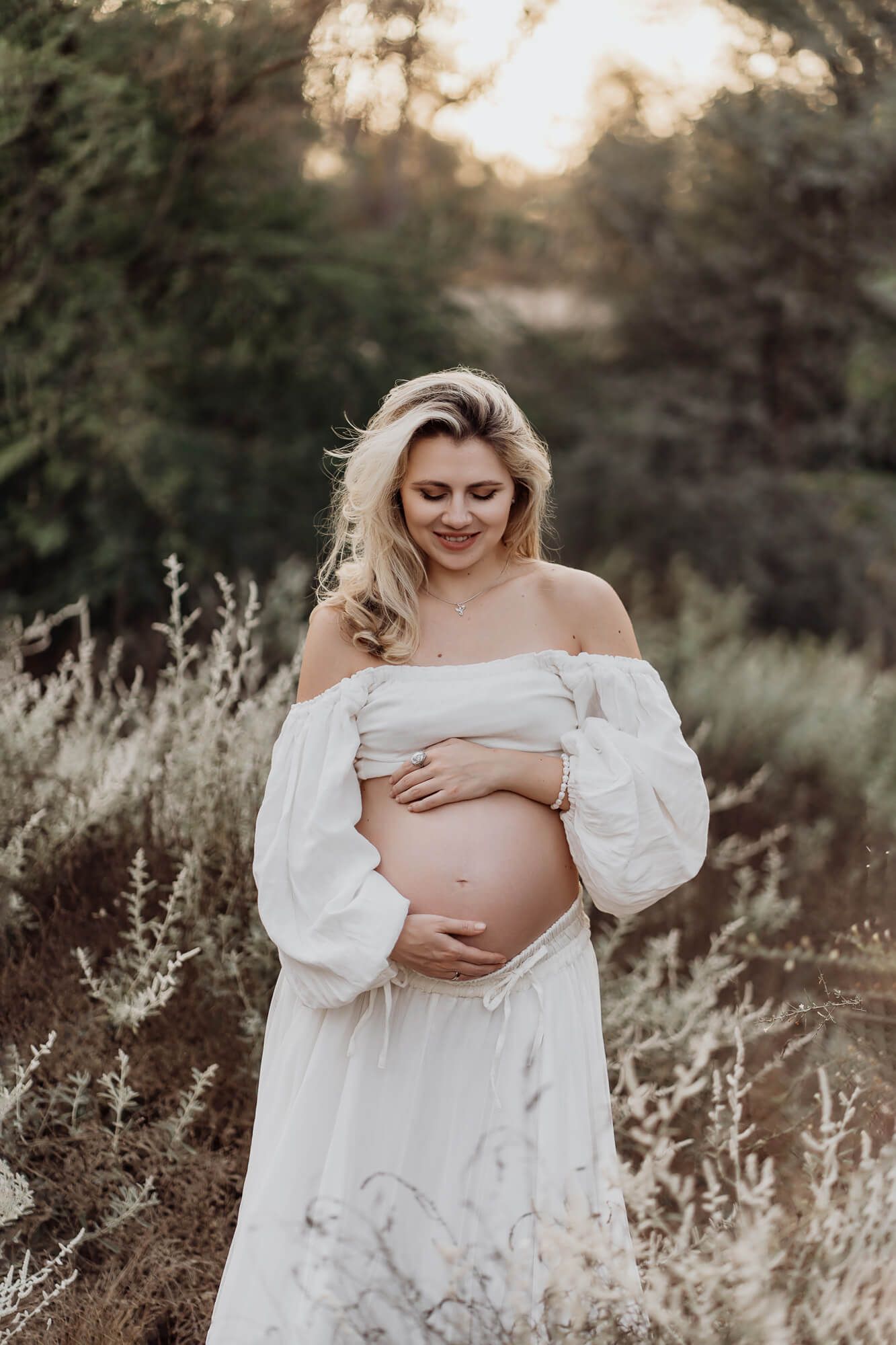 An expectant mother in flowing white dress poses during golden hour in nature.