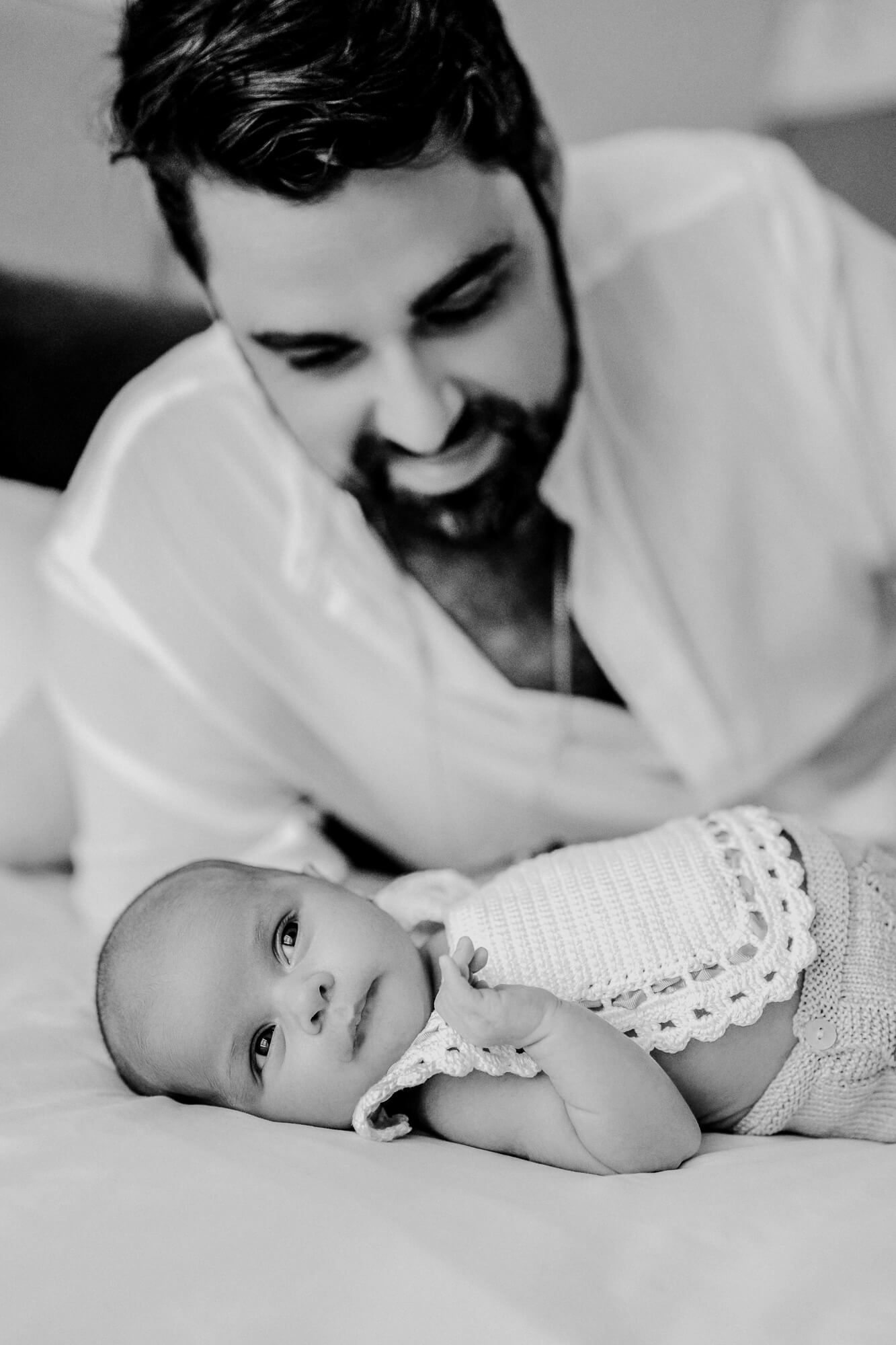 Black and white photo of a tender moment between an adult and a baby in white clothing lying on a bed.