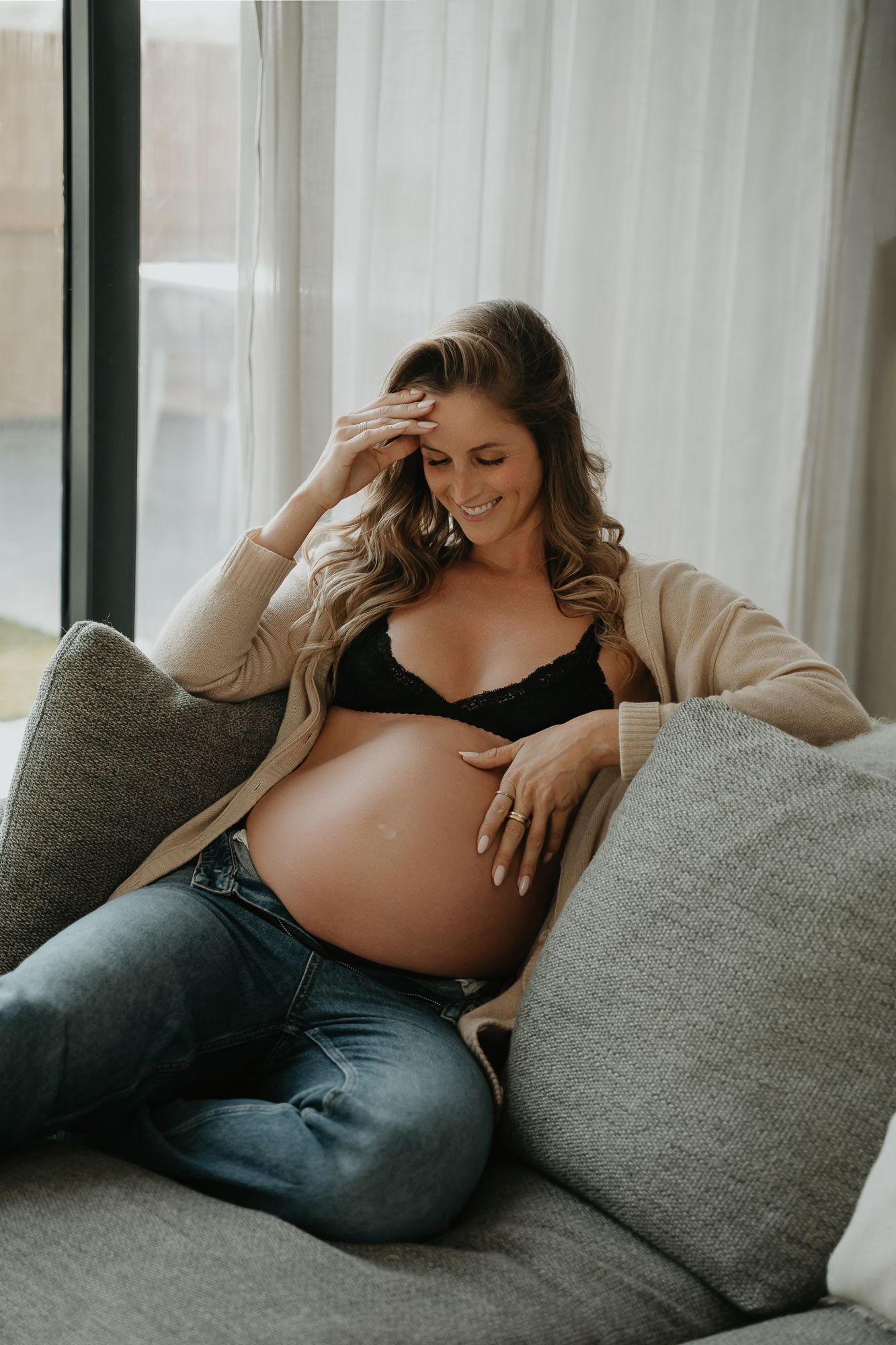 Expectant mother in cozy cardigan and jeans relaxes on grey couch by window with soft natural light.