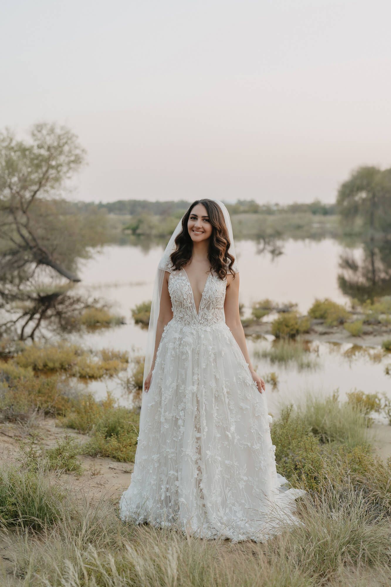 A beautiful white wedding dress with lace details flows elegantly against a lakeside backdrop at sunset.