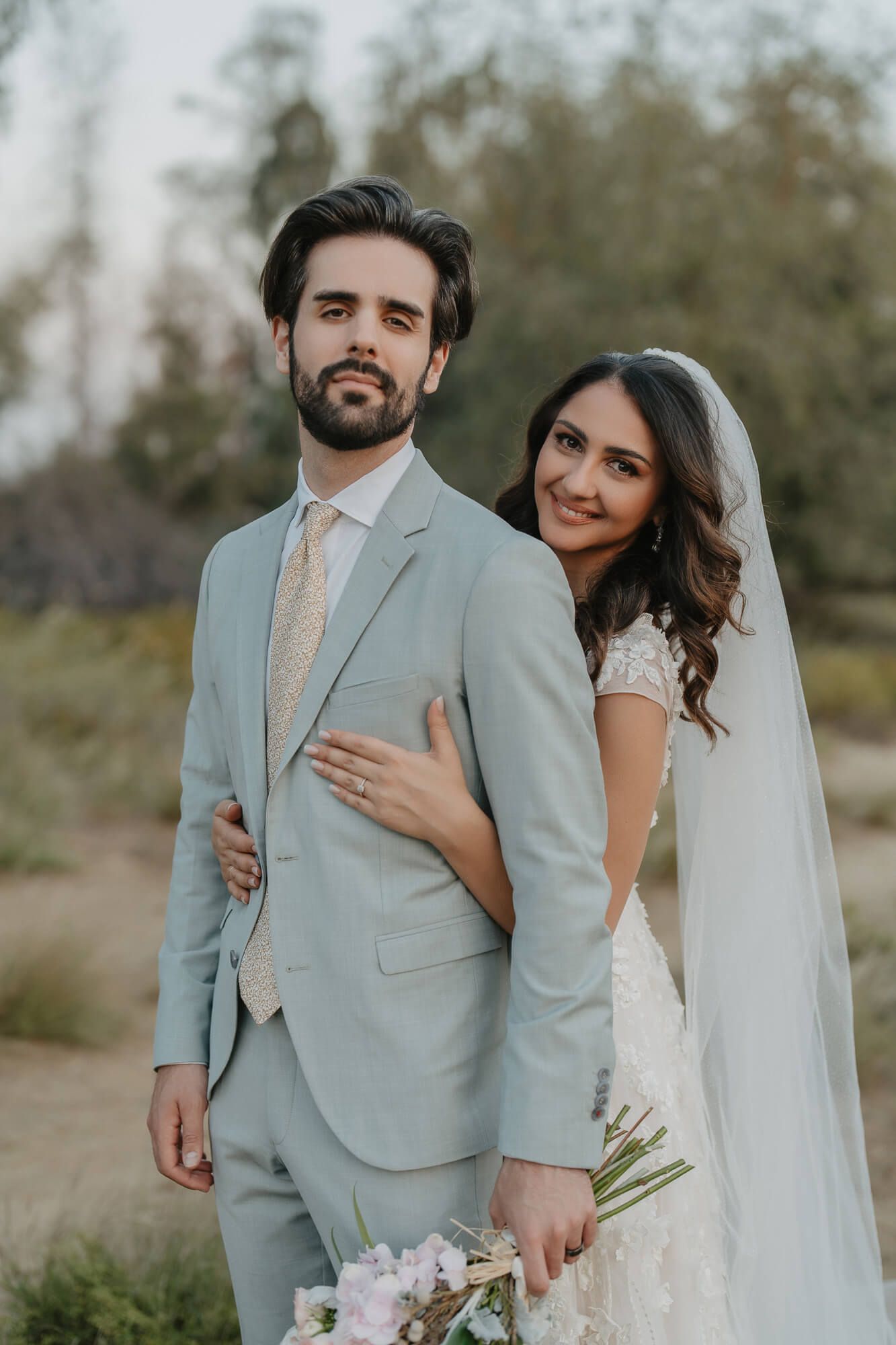 Elegant bride and groom pose together outdoors in a light blue suit and white wedding gown with a flowing veil.