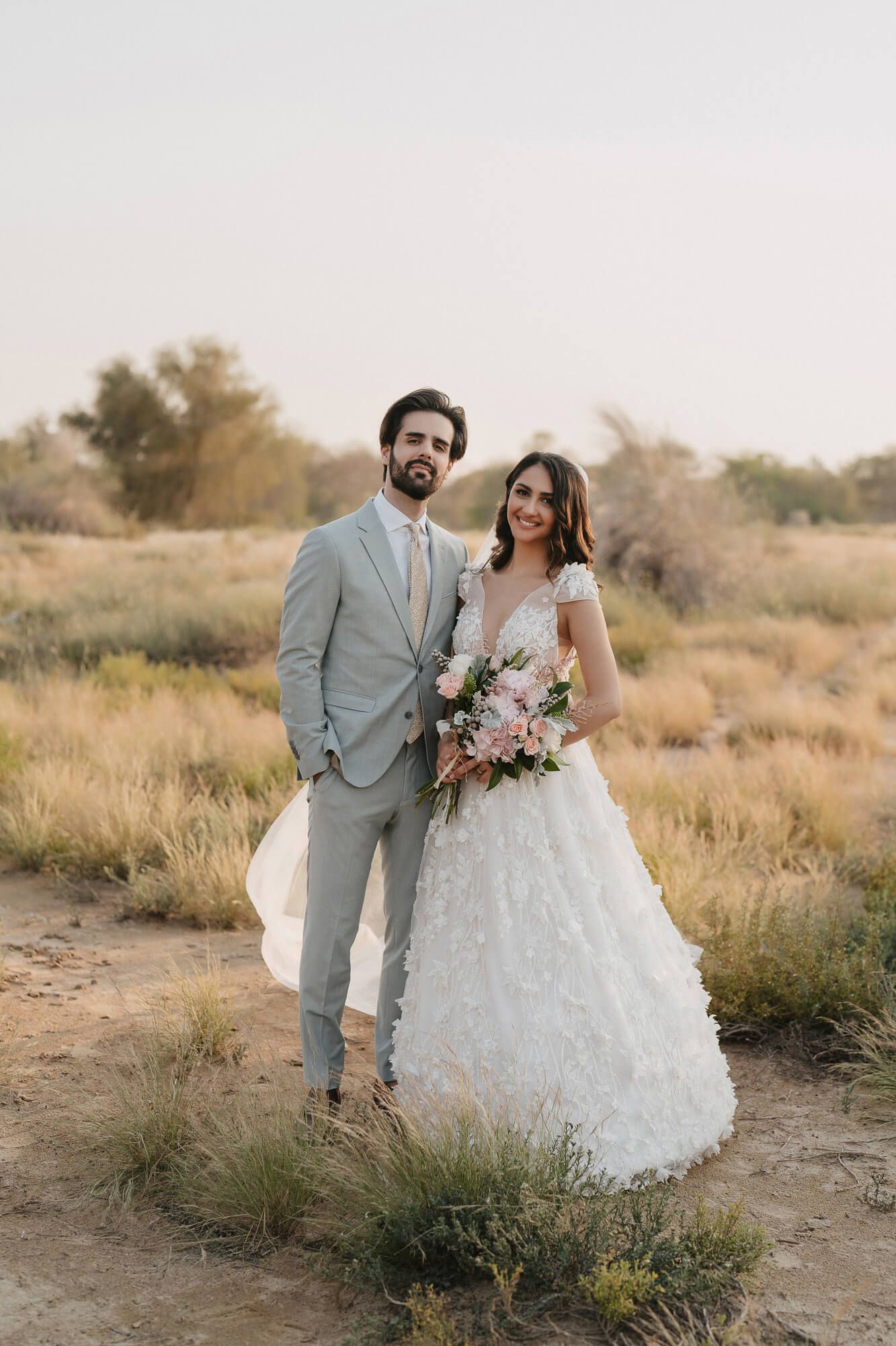 A romantic wedding couple poses in a desert landscape with golden grass and soft lighting at sunset.