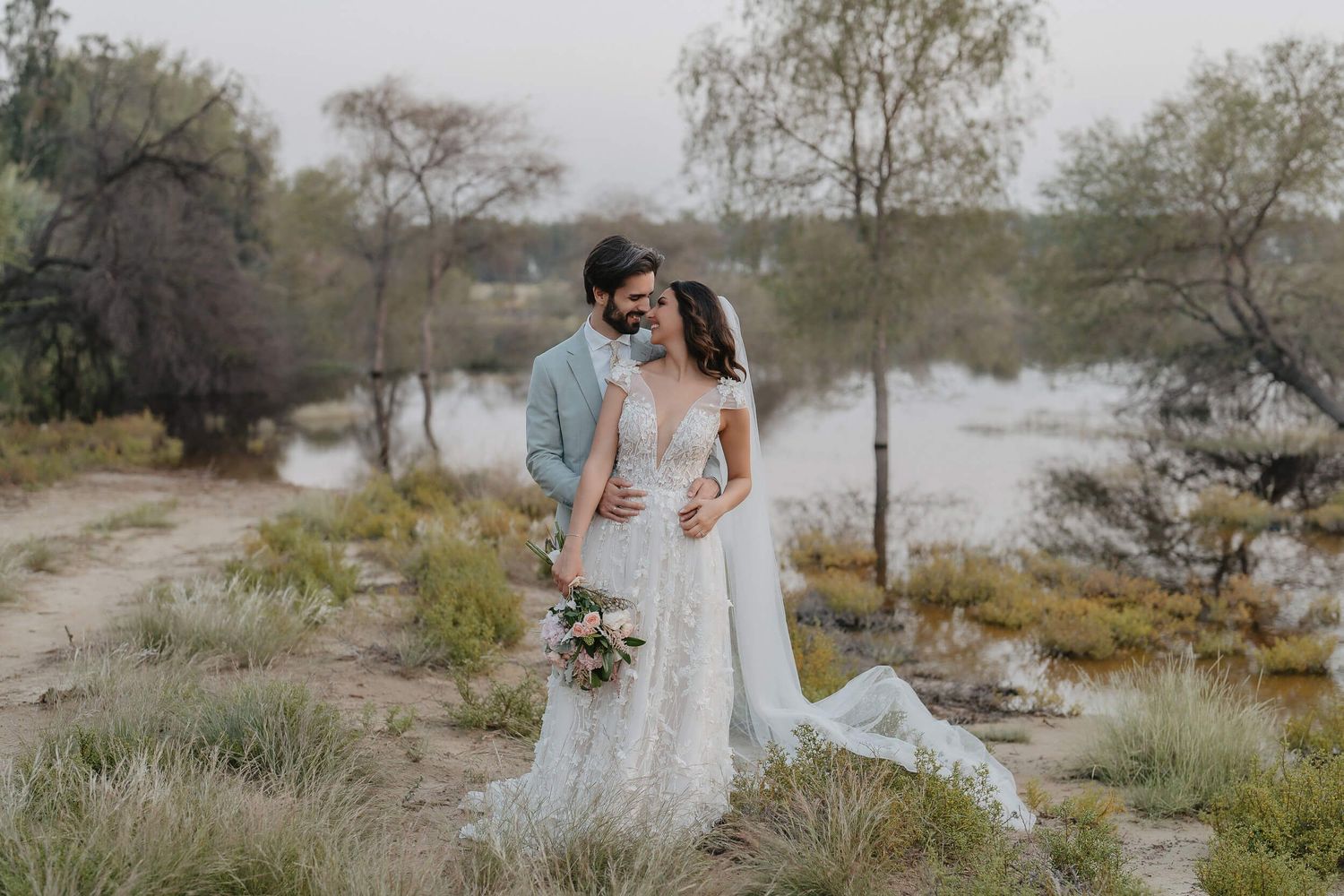 A romantic wedding couple embraces by a misty lake surrounded by bare trees on an overcast day.
