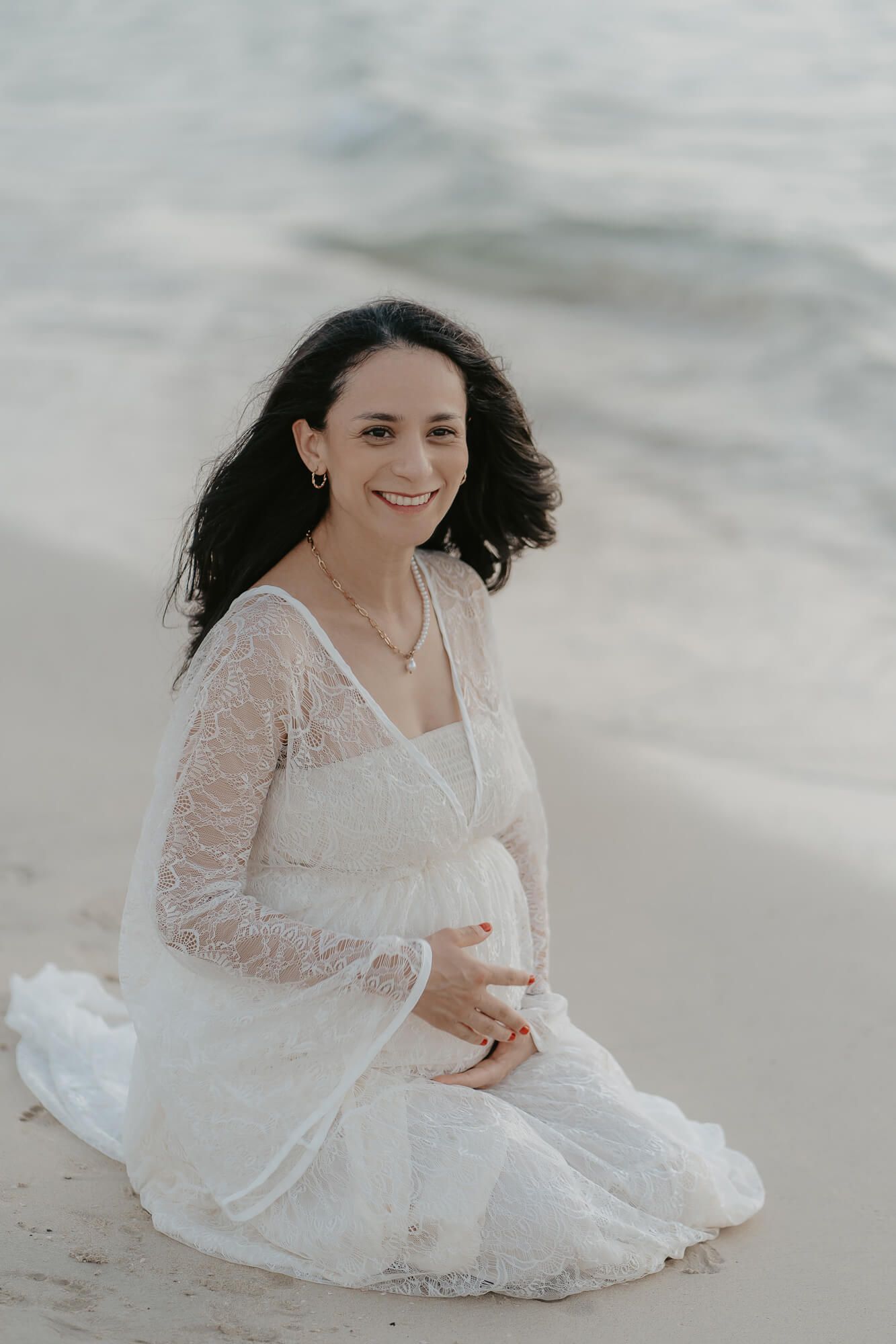 Woman in flowing white lace dress sits gracefully on sandy beach with waves in background.