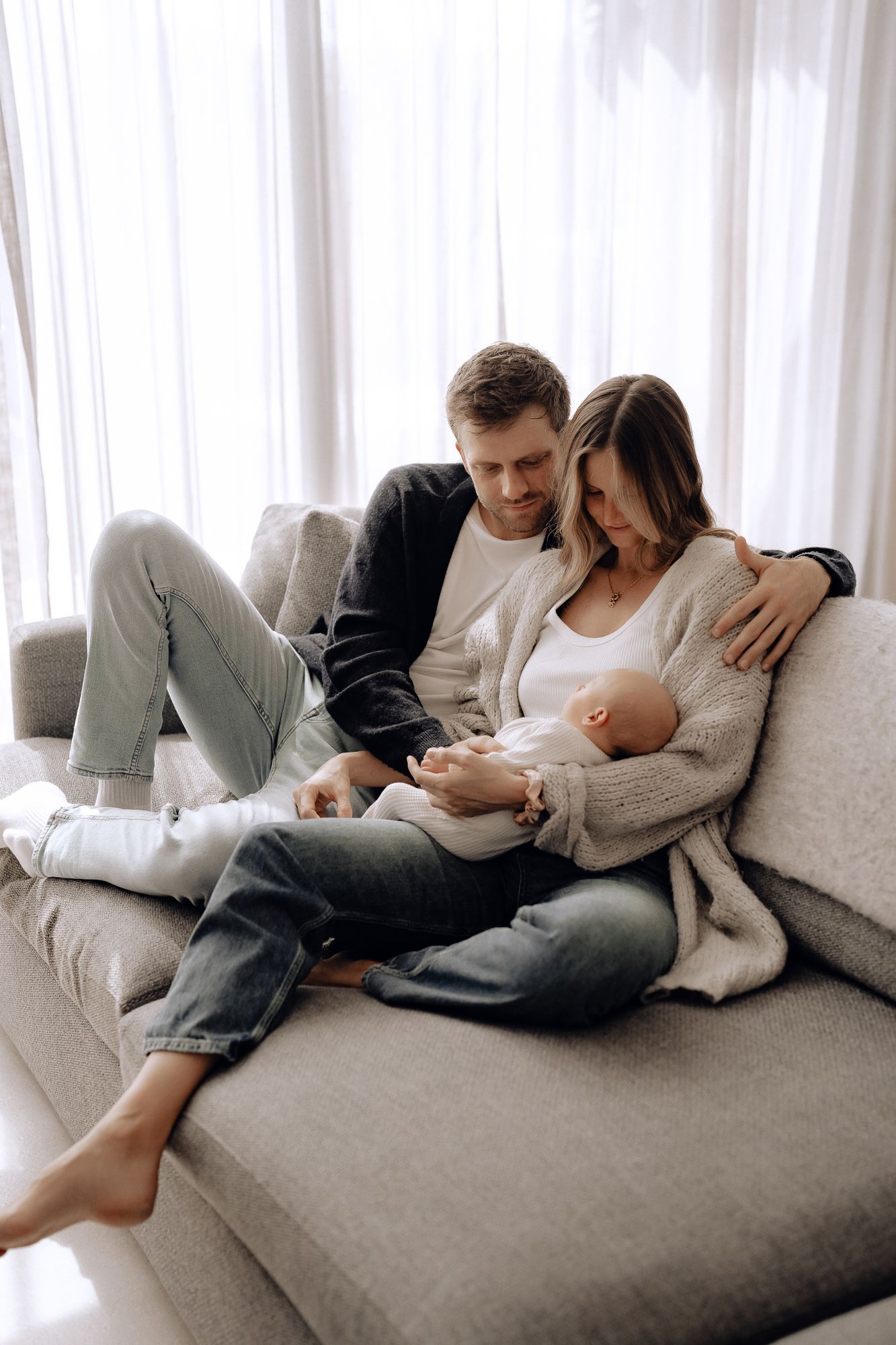 A loving couple relaxes on a gray couch with their newborn baby in a bright, cozy living room setting.