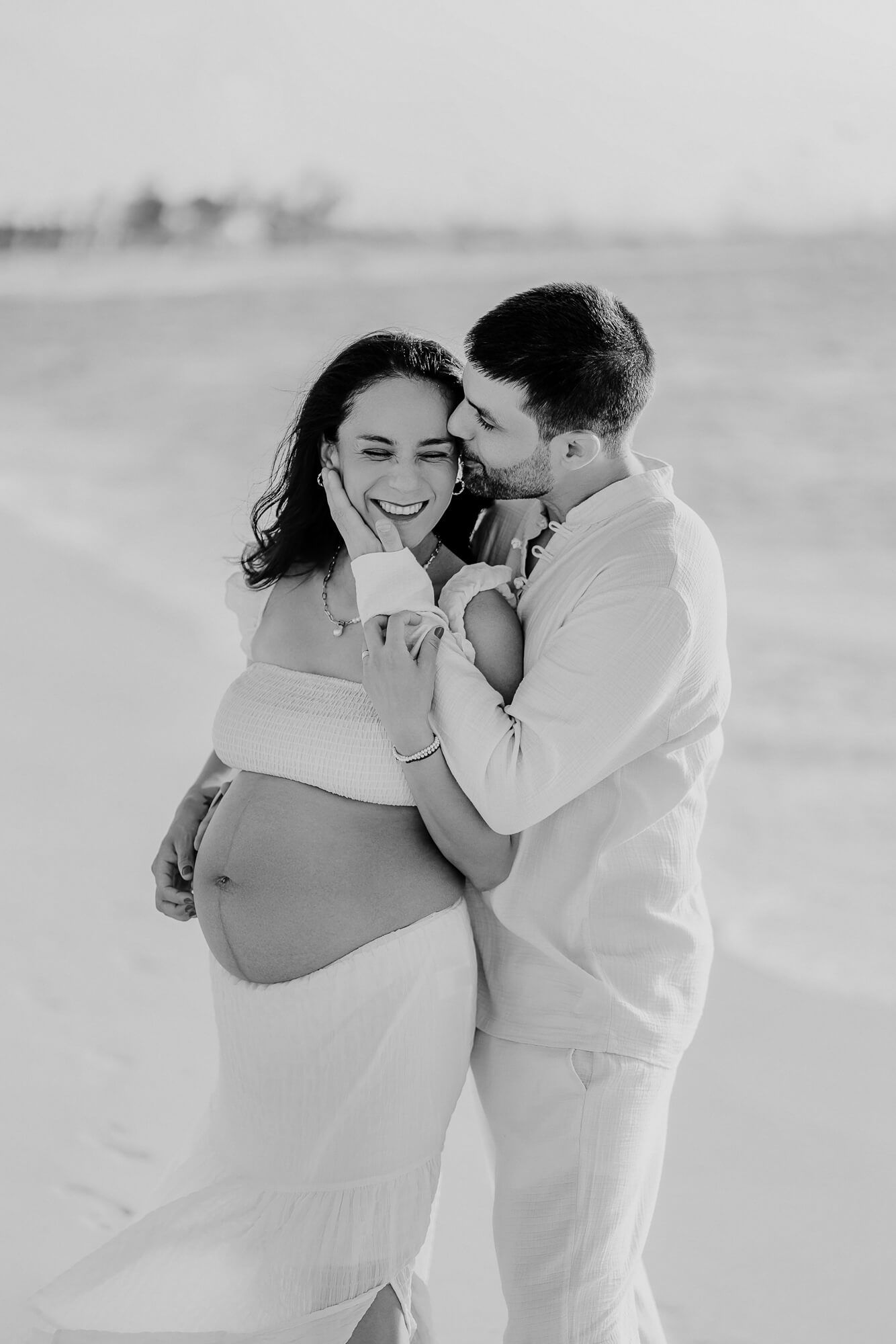 Black and white maternity beach portrait of couple in light colored clothing sharing an intimate moment.