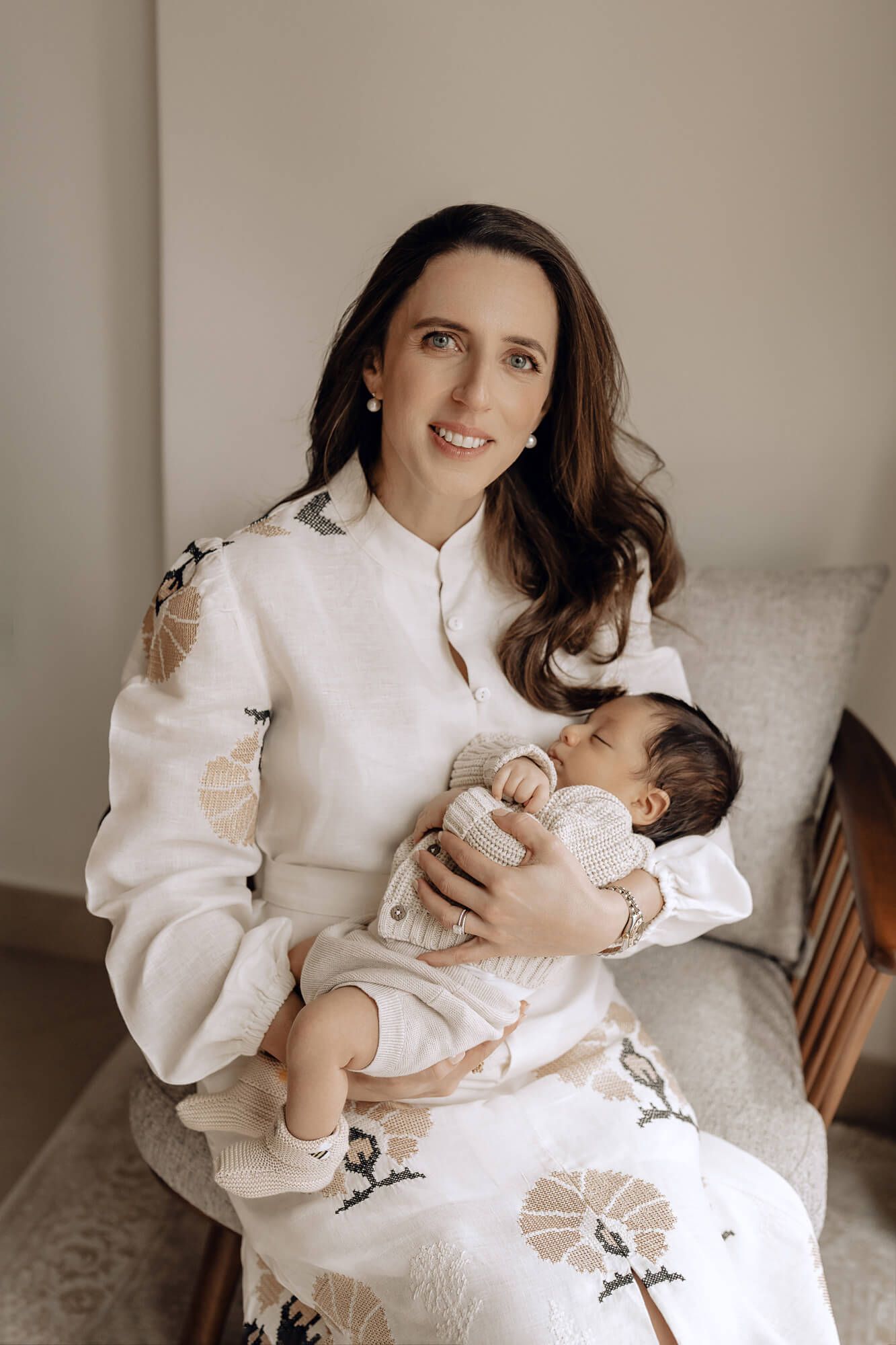 Woman in white sweater holding sleeping baby while sitting on patterned chair.