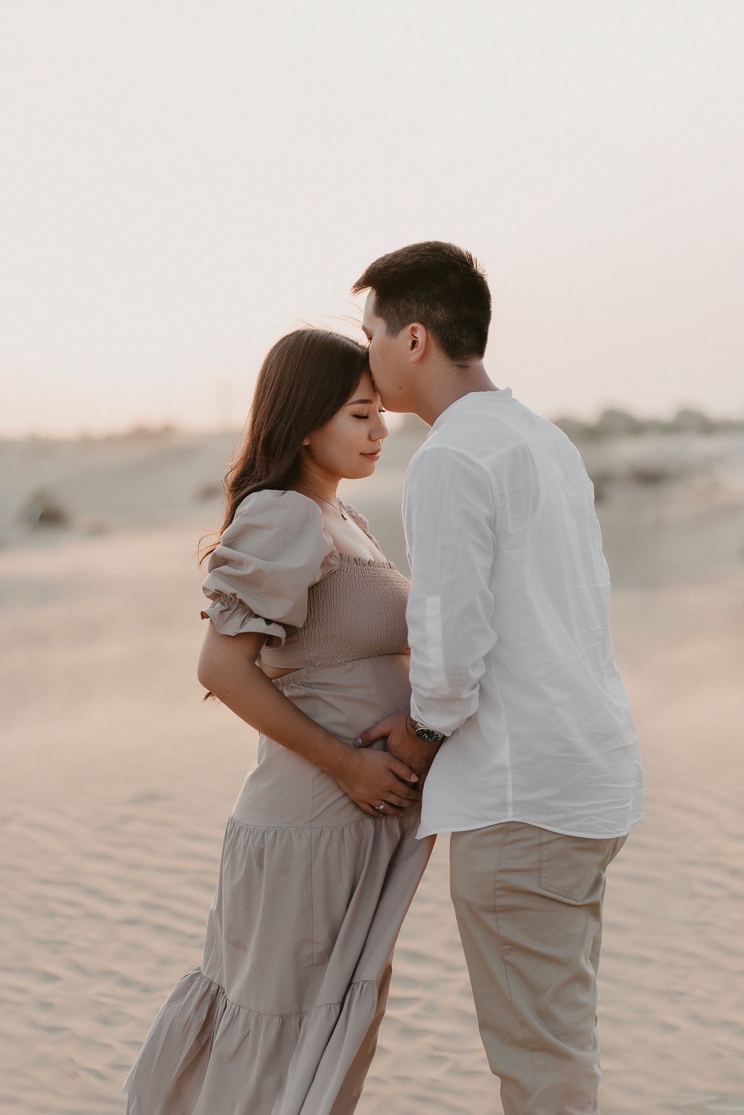 Romantic couple in white and beige outfits embracing tenderly on sand dunes during sunset in desert landscape.