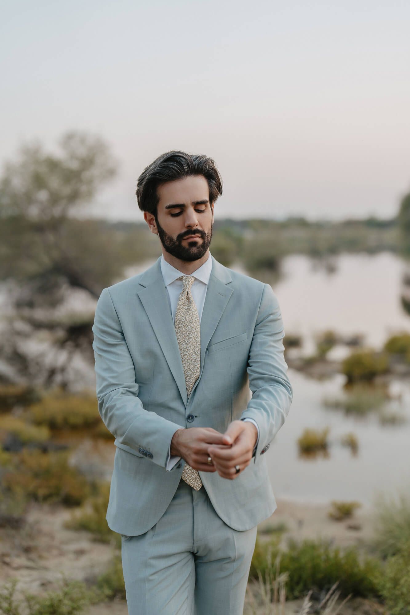 Wedding groom in powder blue suit stands by lakeside on sunny afternoon.