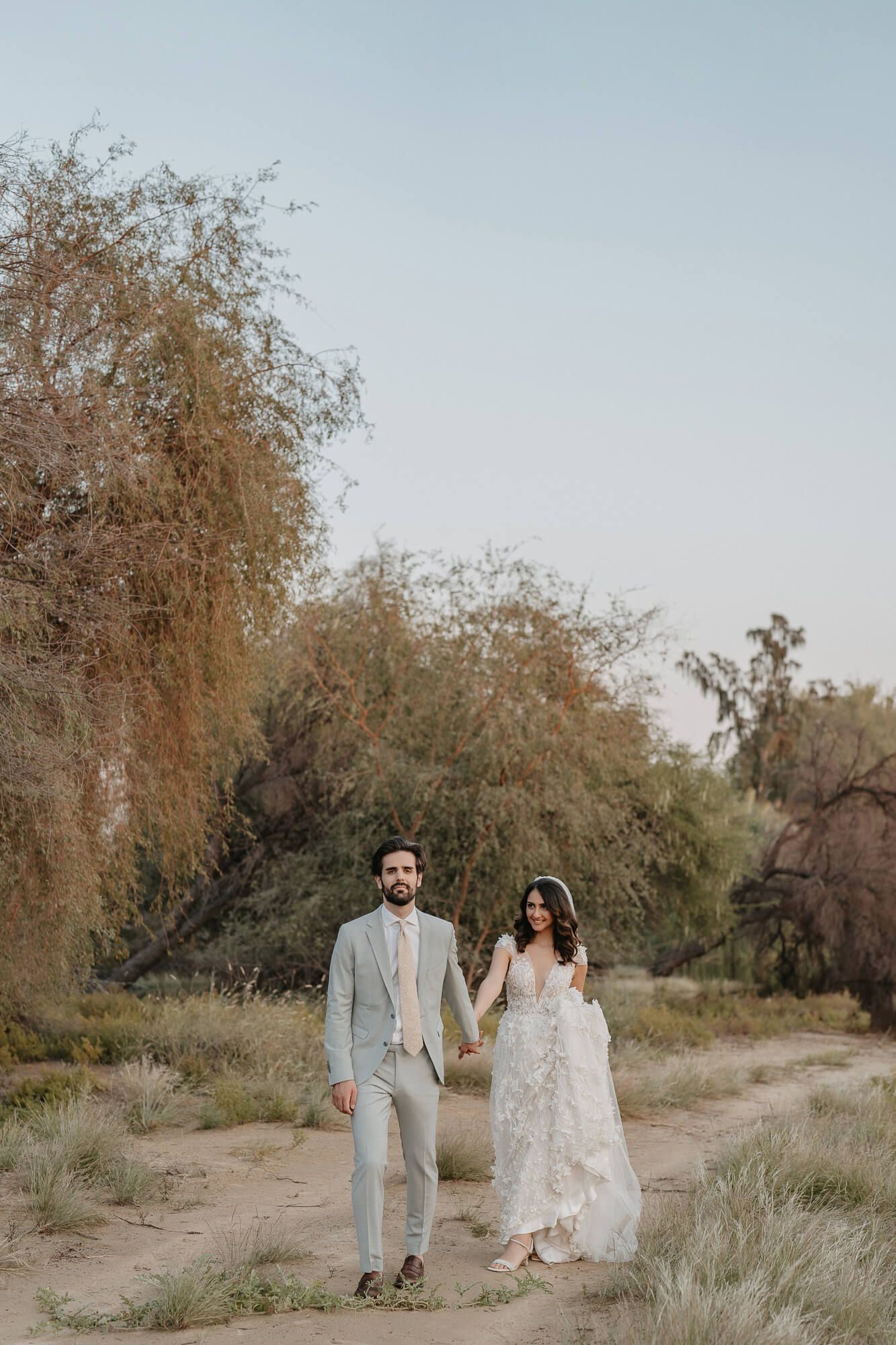 A couple in wedding attire walks along a dusty desert path lined with trees in warm evening light.
