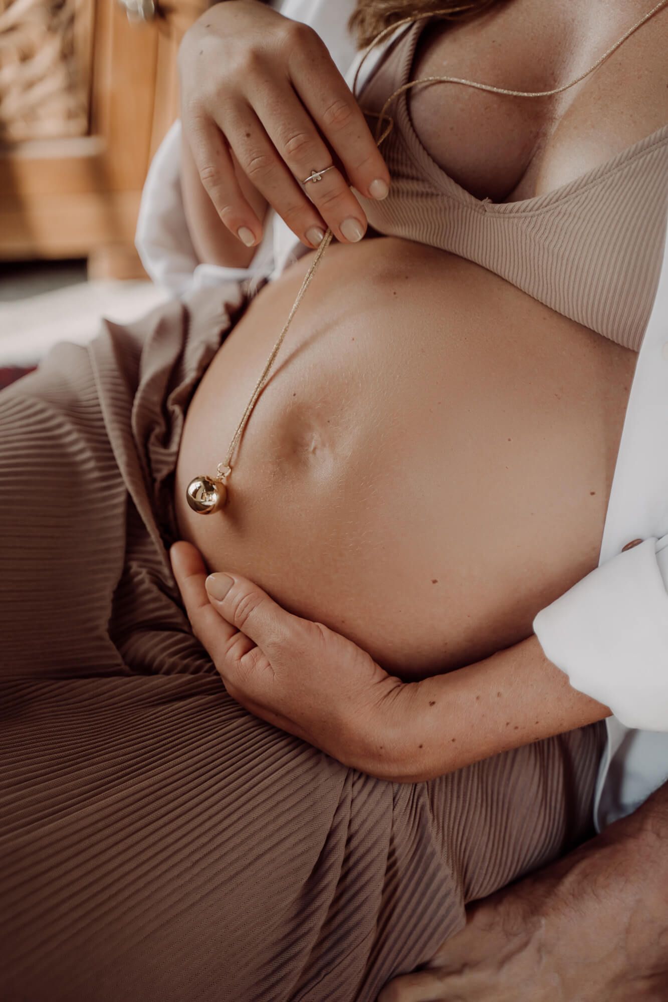 Close-up of pregnant belly with a small gold necklace pendant resting on bare skin in soft warm lighting.