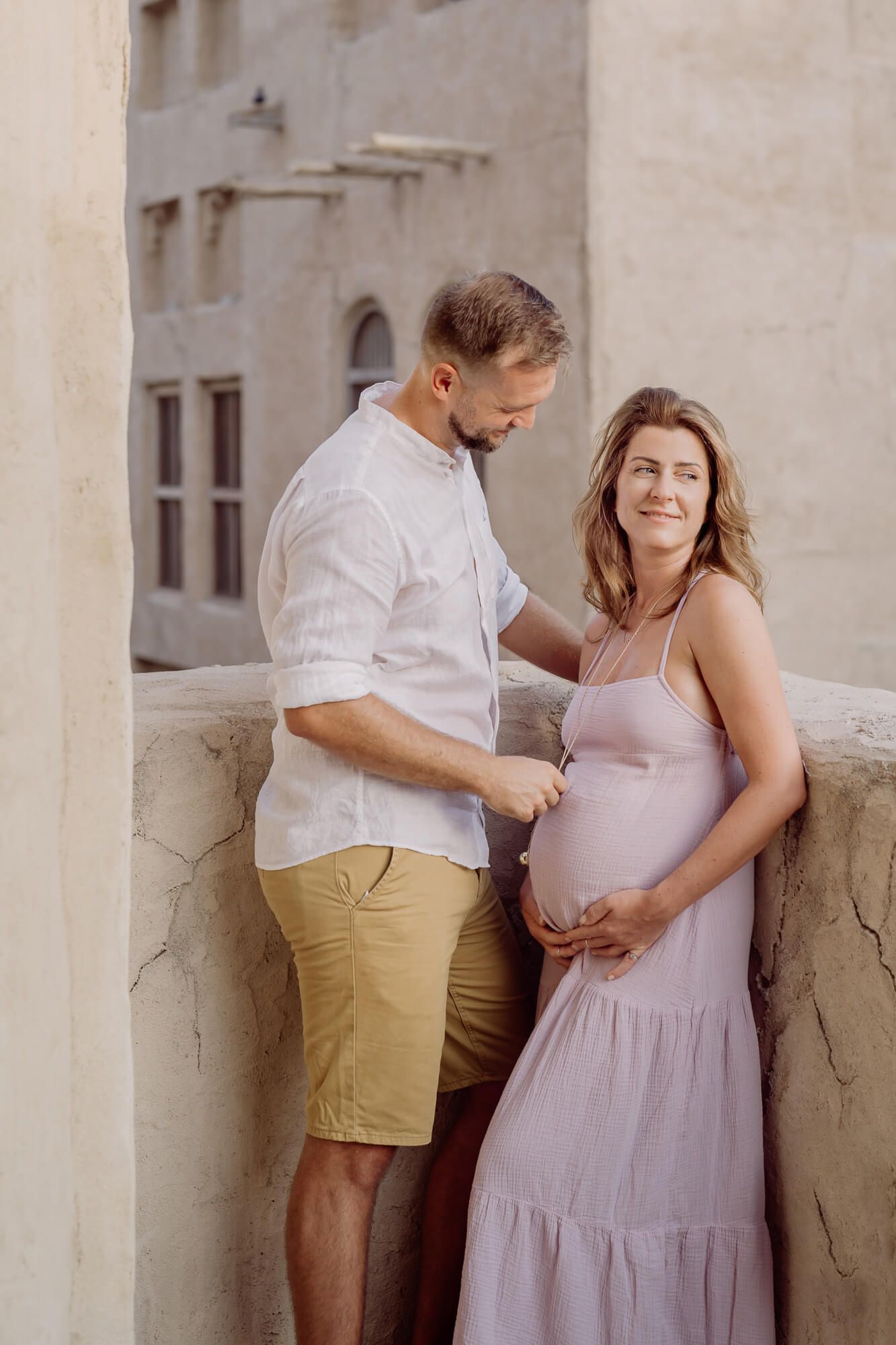 A couple in soft pastels shares intimate moments during a maternity photo session against a neutral wall backdrop.