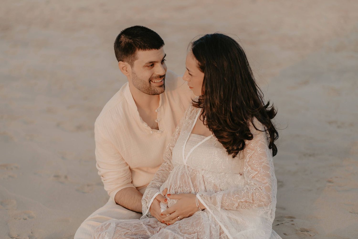 Romantic couple in coordinating white outfits embrace on beach during maternity photoshoot at sunset.
