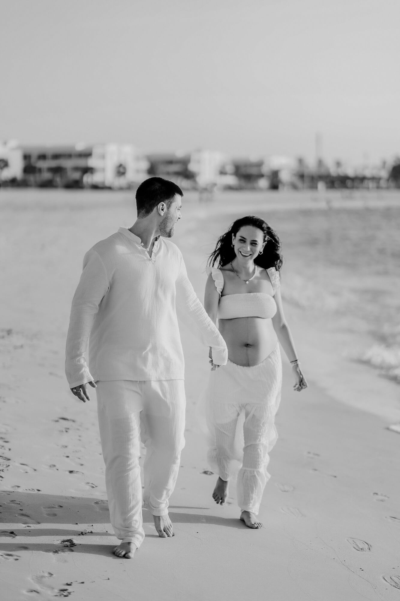 A couple dressed in white outfits walks together along a sandy beach in black and white photography.