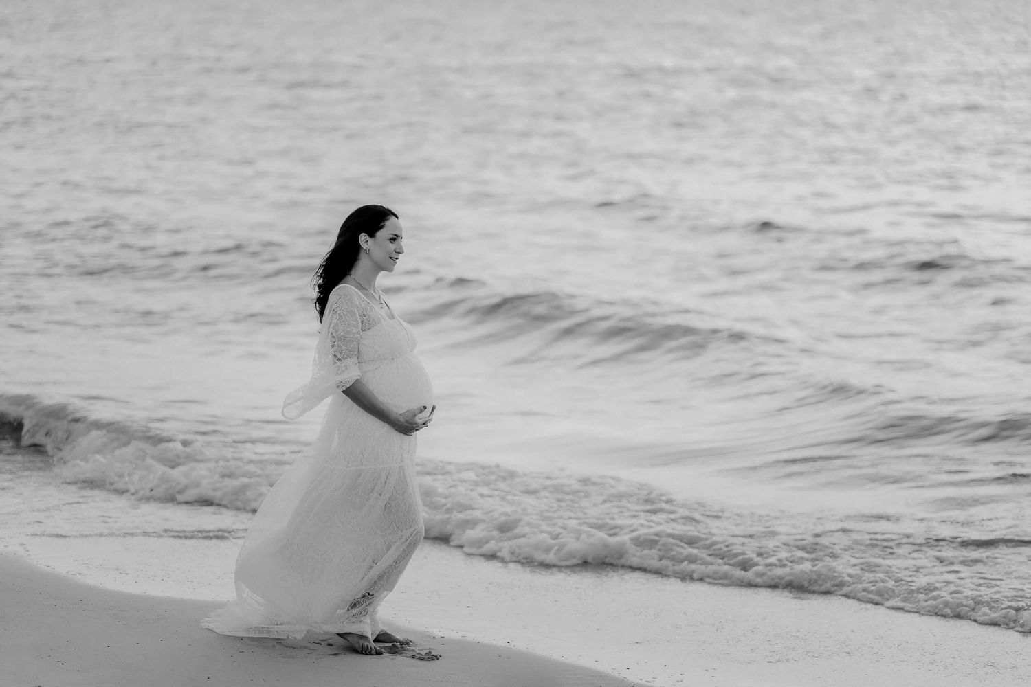 A dreamy black and white beach scene shows a pregnant woman in a flowing white dress against ocean waves.
