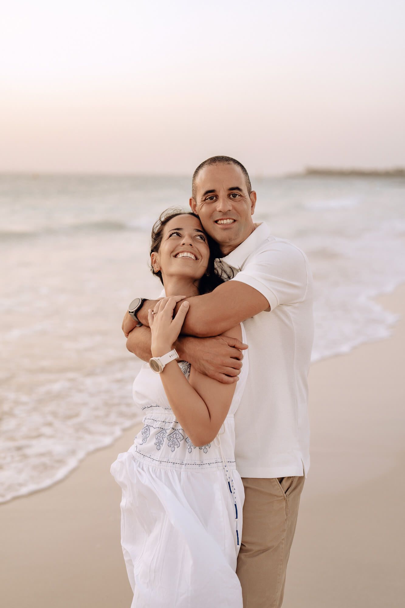 Loving embrace between couple in coordinated white outfits on sandy beach at sunset.